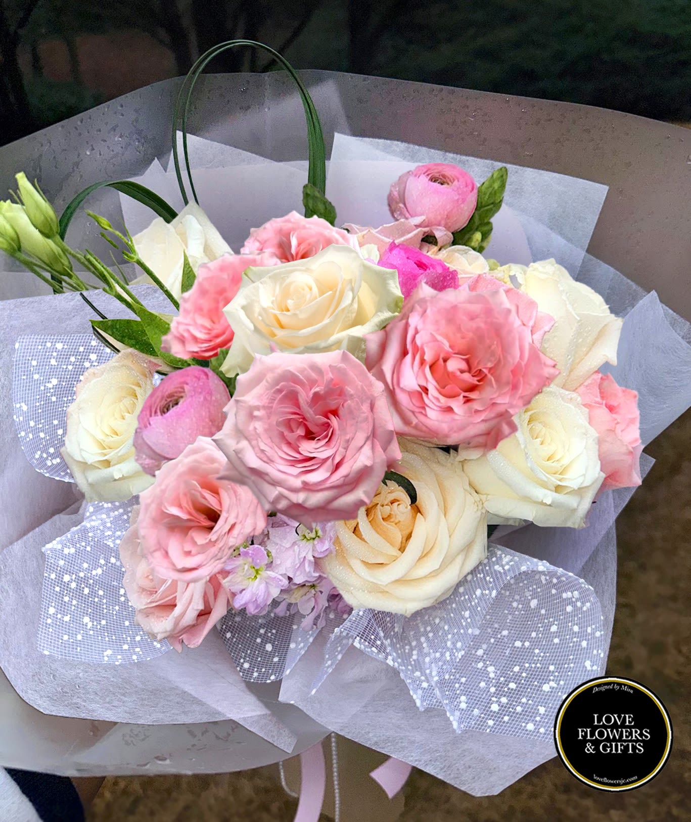 Hand-tied bouquet of pink and cream roses with ranunculus wrapped in white and dotted mesh paper