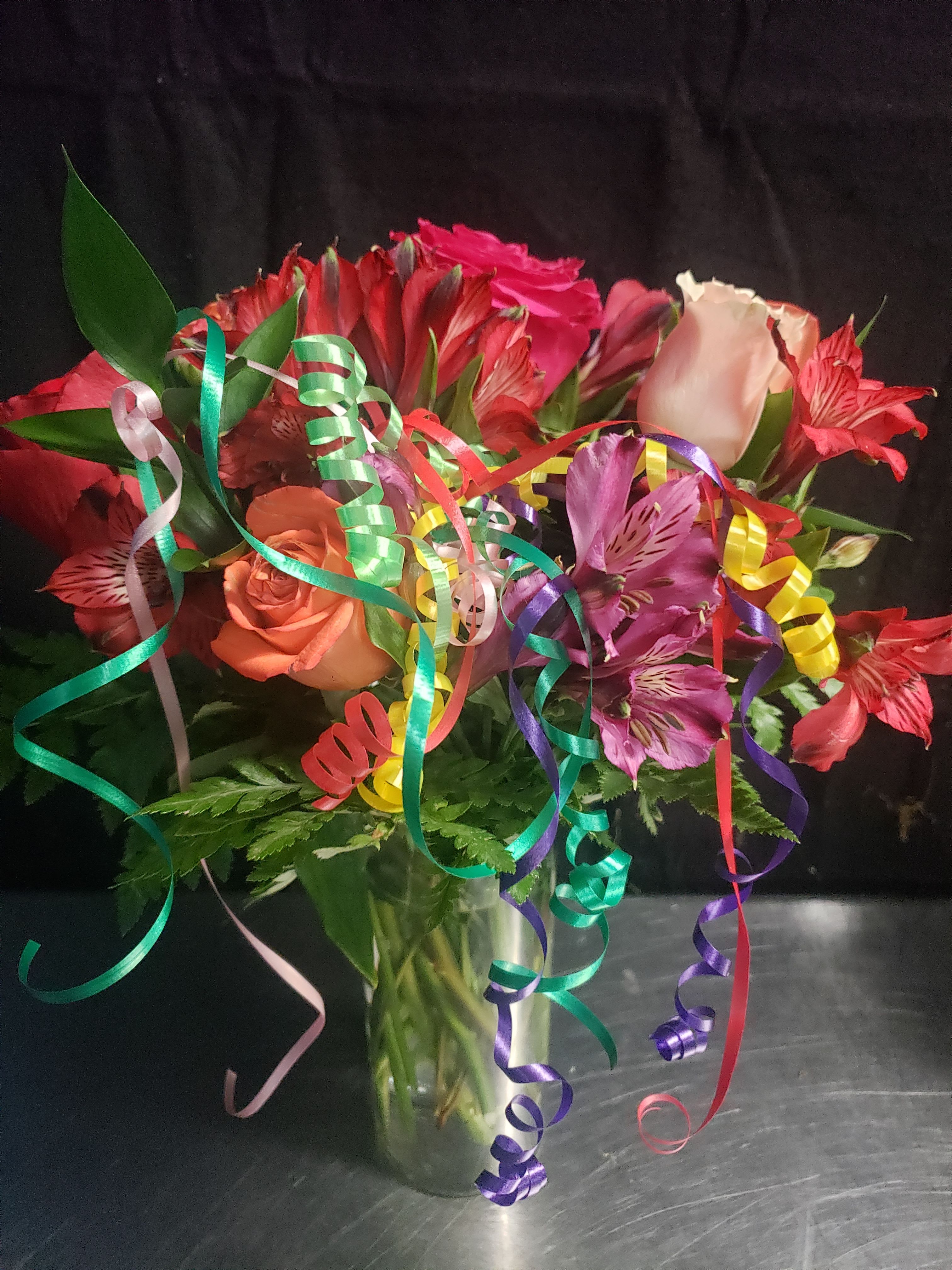 BIRTHDAY BOUQUET - A joyful burst of color that feels like a party in a vase, this birthday bouquet is designed to make the person you're celebrating smile the moment it arrives. Bright pink and red roses mingle with magenta and coral Alstroemeria to create a cheerful, romantic arrangement perfect for birthdays, anniversaries, or any celebratory milestone - the kind of gift that says "you're special" with color and texture.  The arrangement is presented in a clear glass vase with a full, layered composition: tightly clustered roses and alstroemeria create a lush dome of pink, red, purple, and white tones while bright curling ribbons in green, purple, yellow, red, and pink are woven through the stems for playful movement. The glass vase showcases clean lines and lets the blooms take center stage; decorative curling ribbons add festive flair without overwhelming the flowers.  This bouquet suits someone who loves vibrant, feminine color and a lively presentation-perfect for a close friend, partner, or family member who deserves a celebratory surprise. Vase-ready and arranged for immediate display, it arrives ready to enjoy; refresh water daily and trim stems if you'd like to extend the life of the blooms.