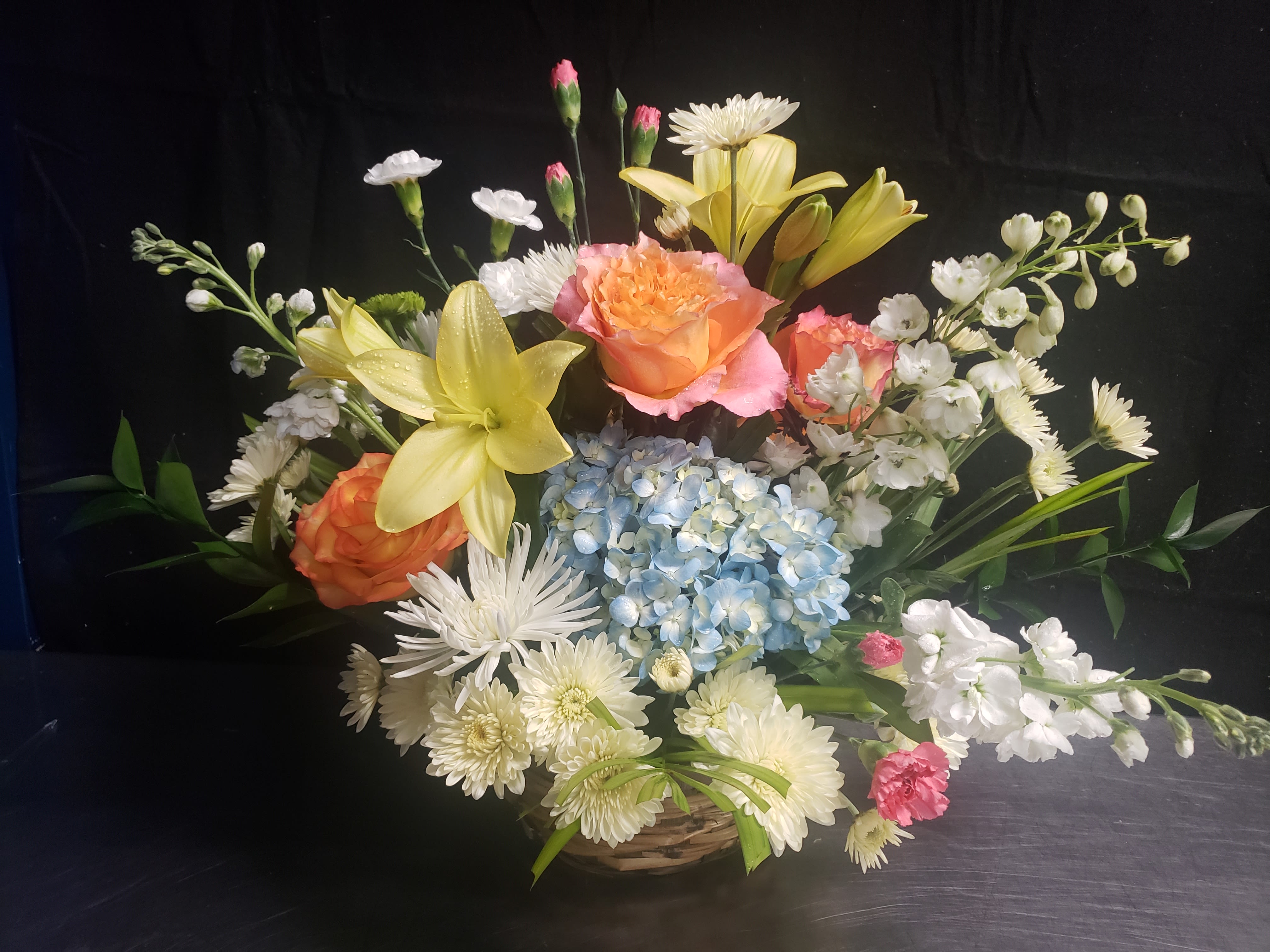 CLEAR SKIES - A lush cluster of blue hydrangeas anchors this event arrangement, surrounded by peach and pink roses, pale yellow lilies, and a halo of white daisy-like blooms-an ideal floral piece for a wedding centerpiece, anniversary celebration, or a tasteful memorial display. The design reads as both cheerful and refined: soft pastels warmed by peach roses and brightened by blue accents, so the person you're honoring will feel both celebrated and comforted.  Arranged in a low woven basket, the composition is full and layered with airy stems and spray flowers that extend outward in a garden-style silhouette. Textures play across rounded hydrangea heads, velvety roses, spiky chrysanthemums and lilies, and delicate daisies for a balanced, formal presentation. The basket makes it easy to place on tables or display areas and gives the arrangement a rustic, gift-ready finish.