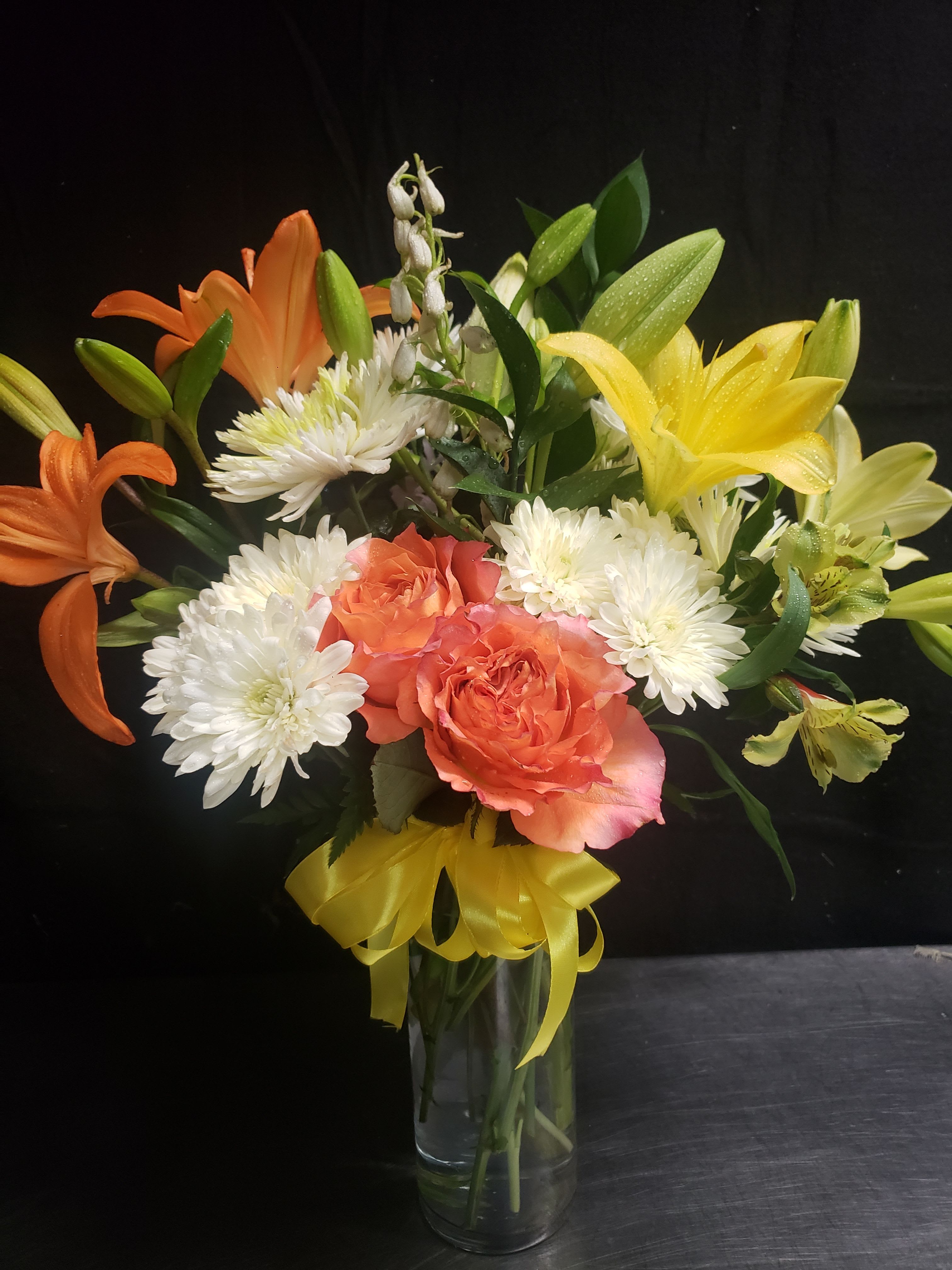 LOVELY BPUQUET - Bright citrus tones of orange roses and yellow lilies make this flower arrangement a cheerful birthday bouquet or congratulations gift that will make the person you're thinking of feel celebrated. The lively mix of orange, white, yellow, and green reads joyful and fresh-perfect for springtime surprises, thank-you gestures, or anytime you want to brighten someone's day.  This full, rounded arrangement pairs orange roses with open yellow-and-orange lilies, white daisy-like chrysanthemums, pale alstroemeria and daisies for soft contrast. The stems are gathered in a clear glass vase and finished with a bright yellow ribbon tied at the neck, giving it a gift-ready presentation. The saturated colors and varied textures-velvety rose petals, starry lilies, and daisy centers-create a warm, uplifting display that arrives ready to enjoy.