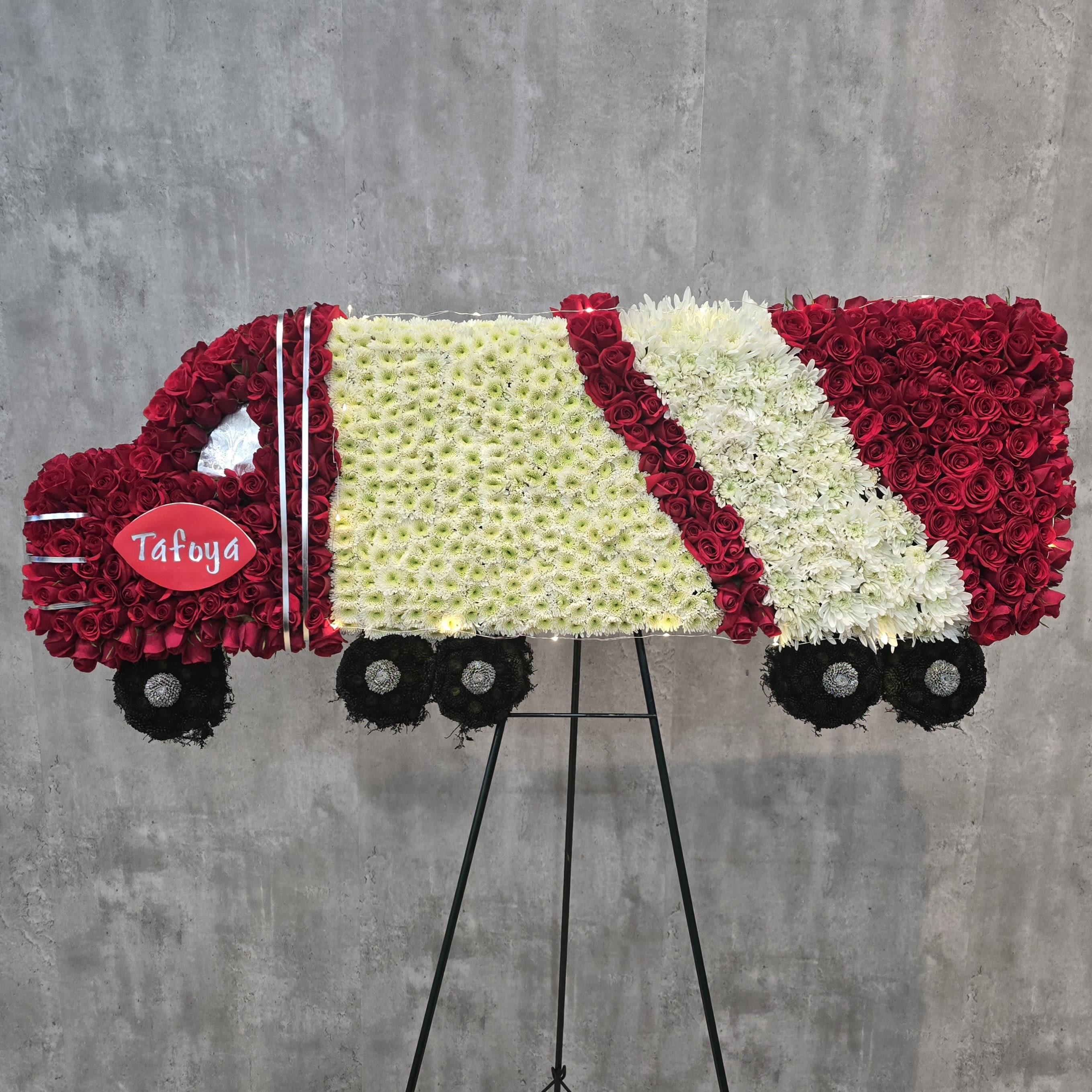 Semi Truck Tribute - A handcrafted semi truck tribute covered in white chrysanthemums with red accents creates a memorable sympathy and funeral arrangement for drivers, fleet workers, or anyone who loved the open road. This semi truck floral tribute reads clearly from a distance-playful yet respectful-so the person you're honoring will feel seen and celebrated for the miles they logged. Use this truck floral display as a standout memorial at a funeral, graveside service, or celebration of life.  The large floral display is shaped like a truck and measures about 32" long by 12" tall, mostly covered in white mums with red roses  defining the cab, tinted wheels, and metal trim. Black floral detailing marks the tires and outline, while mixed bouquets and seasonal filler flowers add texture and contrast. A decorative truck sculpture anchors the piece, a string of small lights is woven through the flowers for a gentle glow, and it arrives ready to stand on the included metal easel.  Ideal as a semi truck floral tribute or novelty truck arrangement for memorial services, this piece speaks directly to someone who lived for the road-family members and coworkers will recognize the personal touch. We can discuss custom color swaps or adding a company logo to personalize the tribute; contact our designers to arrange customization or delivery details.