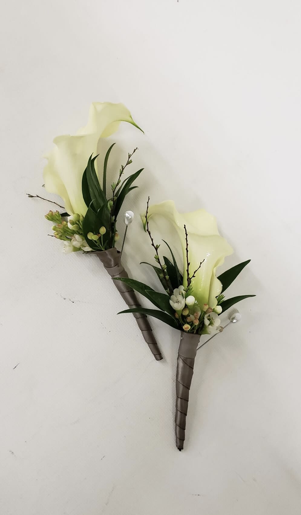 Two white calla lily boutonnieres with dark green leaves and ribbon-wrapped stems