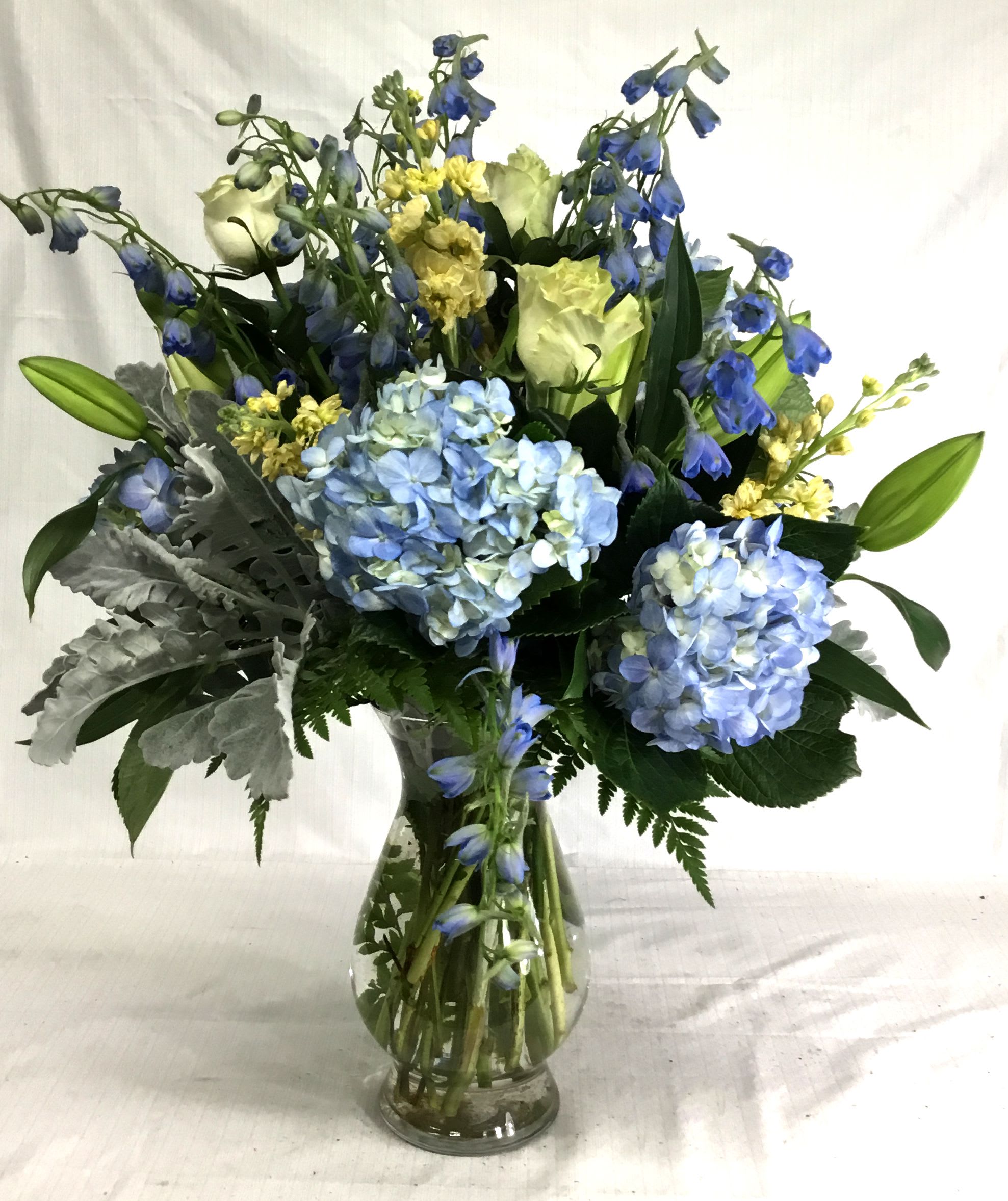 Tall arrangement of blue hydrangeas, white roses, lilies, and yellow flowers in a clear glass vase.