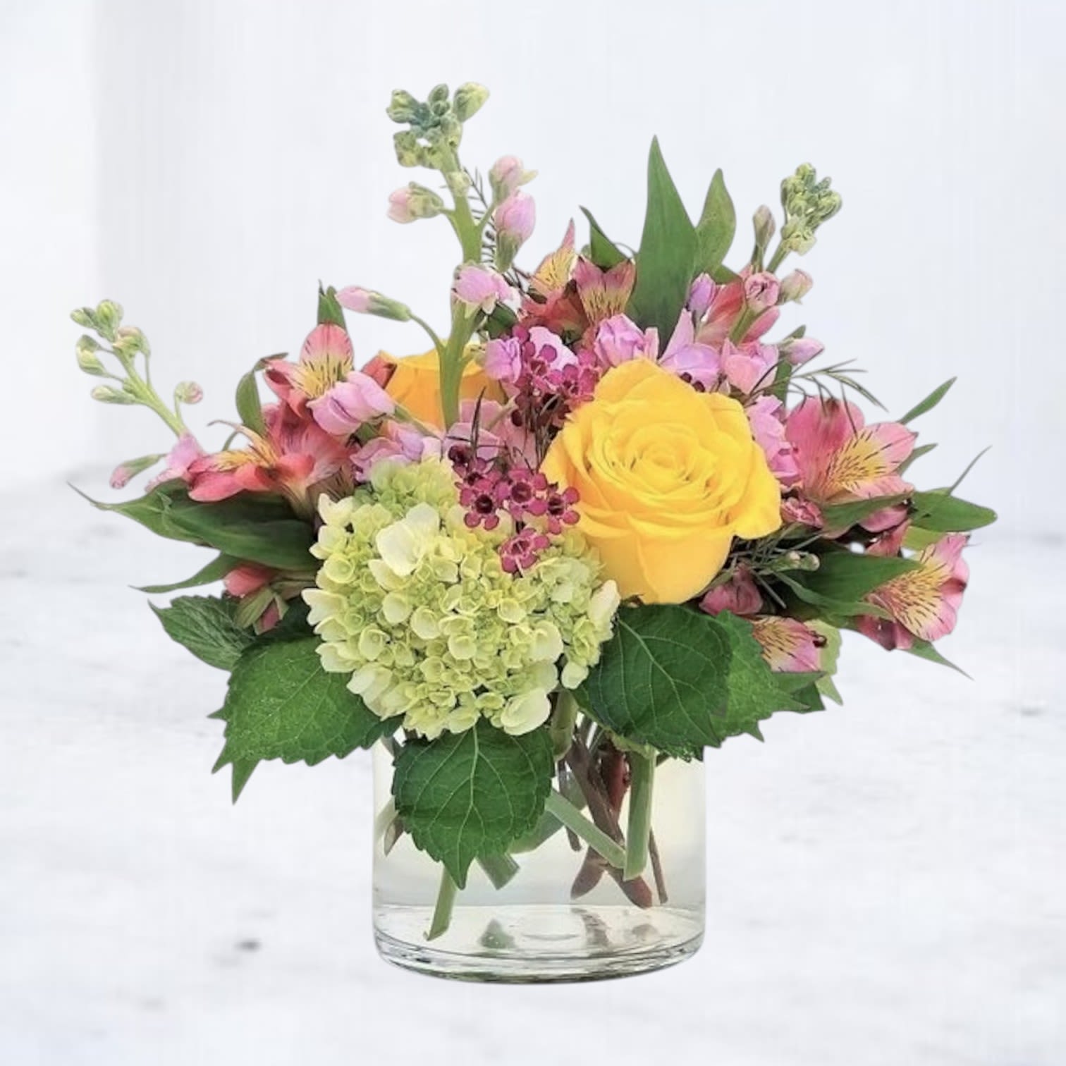 Mixed bouquet of yellow roses, pink flowers, and green hydrangea in a glass vase