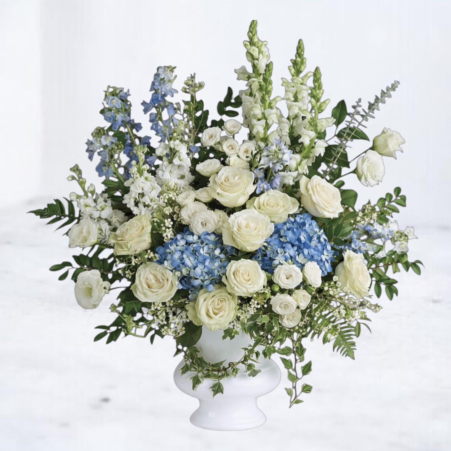 Tall white and blue arrangement with roses and hydrangeas in a white pedestal vase