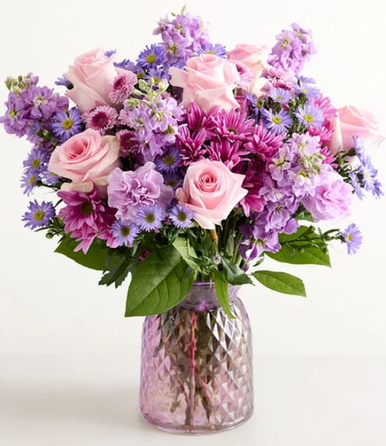 DAY DREAM BOUQUET - Soft pink roses take center stage in this Day Dream bouquet, surrounded by lavender and purple accents for a gentle, romantic birthday or anniversary arrangement. The combination reads as a thoughtful spring celebration gift that will make the person you're thinking of feel cherished and seen-soft, feminine colors that say "I'm celebrating you" without being over the top.  Clusters of chrysanthemum blossoms, dainty aster blooms, and ruffled stock add layered texture and movement around the larger roses, giving the bouquet a full, rounded silhouette. Arranged in a clear faceted glass vase, the mix of pink, purple, and white creates a polished, gift-ready presentation that looks lovely on a mantel, dining table, or bedside.    Perfect for celebrating birthdays, anniversaries, or simply brightening someone's day, this bouquet balances romantic charm with cheerful energy. The glass vase makes it easy to display; change the water every other day and trim stems as needed to help blooms last.