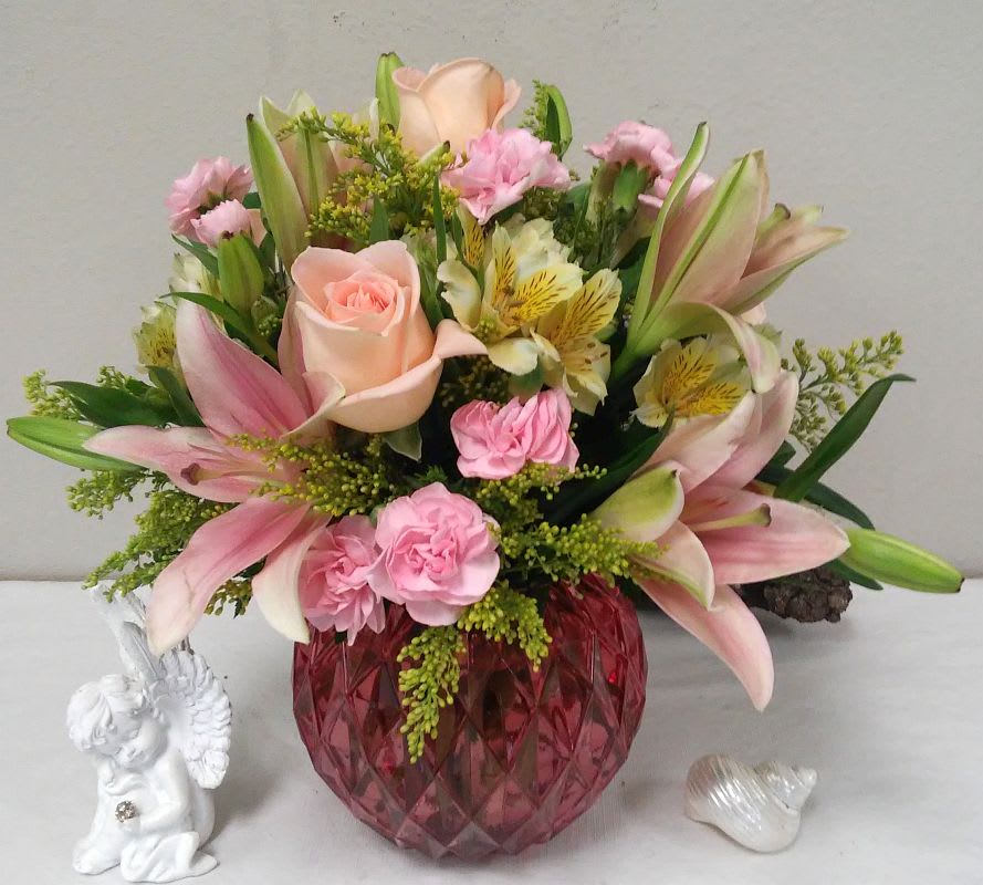 Pink lilies and roses arranged in a red glass vase with a small angel figurine