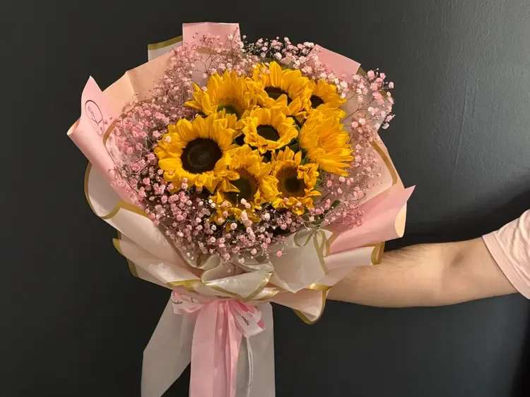 Sunlit Blush - This cheerful bouquet features 10 glowing sunflowers, symbolizing happiness, warmth, and positivity. Each bloom is beautifully framed with airy pink baby’s breath, creating a soft romantic contrast to the bold yellow petals. The combination feels both vibrant and elegant — perfect for birthdays, celebrations, or simply to brighten someone’s day. This design carries a joyful energy while maintaining a refined, modern look.
