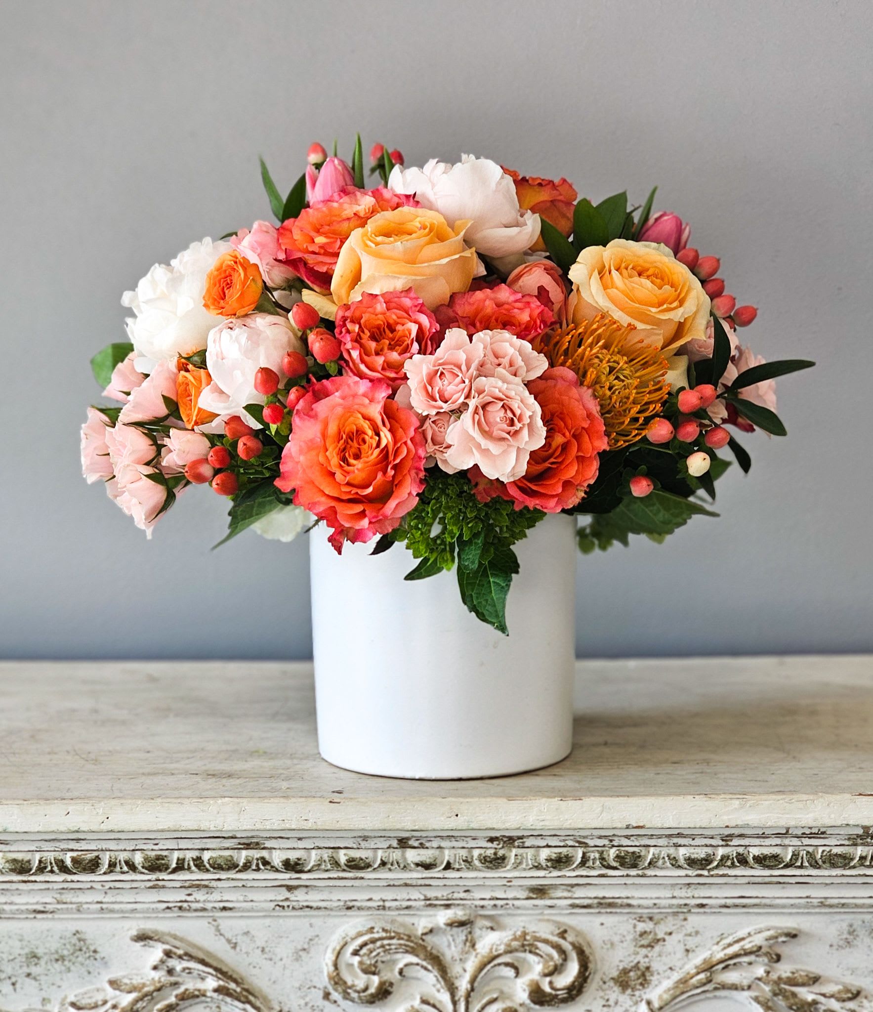 Clementine - A beautiful color palette similar to a sunrise in shades of blush to orange using seasonal premium flowers such as roses, hydrangea and protea in a modern white ceramic vase.