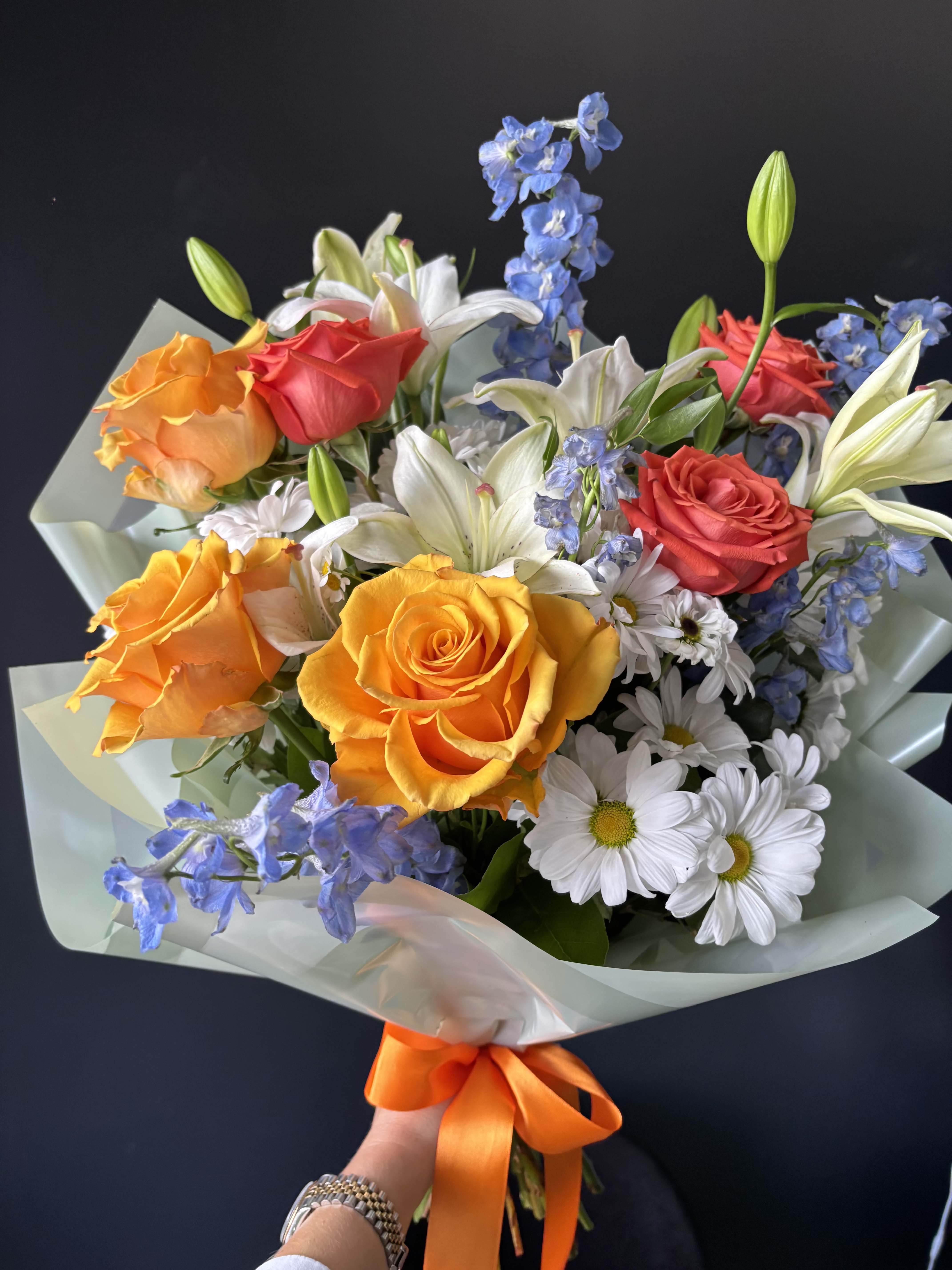 Velvet Sunrise - Velvet Sunrise is a radiant composition inspired by the soft glow of early morning light. Rich orange and coral roses create a warm, velvety focal point, symbolizing energy, joy, and refined beauty. Elegant white lilies add a sense of purity and sophistication, gently opening the bouquet and giving it a graceful structure. Airy blue delphinium introduces contrast and depth, bringing a fresh and modern touch to the arrangement. Delicate daisies soften the look, adding a natural, effortless charm. Wrapped in soft mint-toned paper and finished with a vibrant orange ribbon, this bouquet feels both luxurious and uplifting — perfect for meaningful gifts, celebrations, or simply brightening someone’s day with style.