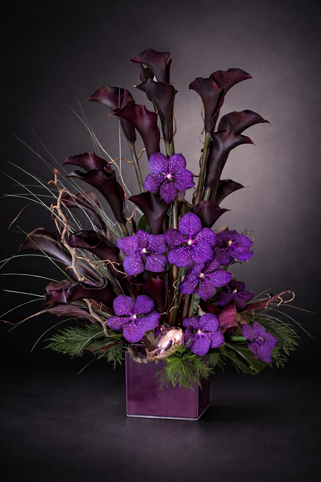 Silence at Night - Tall, dramatic dark purple calla lilies take center stage in this vertical, sculptural flower arrangement-perfect as a  for an elegant evening event. The deep burgundy-black of the callas contrasts strikingly with clusters of vivid violet filler blooms, creating a moody, refined statement that will make the person you're thinking of feel seen and honored.  Set in a clear glass vase, the composition is accented with thin decorative twigs that add spiky, airy texture and a modern architectural silhouette. The arrangement's strong vertical lines and focused color palette of purple, black, and green give it a formal, contemporary presence that works well on a mantel, reception table, or beside a bedside.  Choose this arrangement when you want to convey thoughtful sympathy, quiet strength, or sophisticated style-the kind of bouquet that feels both composed and emotionally resonant. Care is simple: keep water fresh, trim stems every few days, and place away from direct heat to preserve the calla lilies' elegant shape.