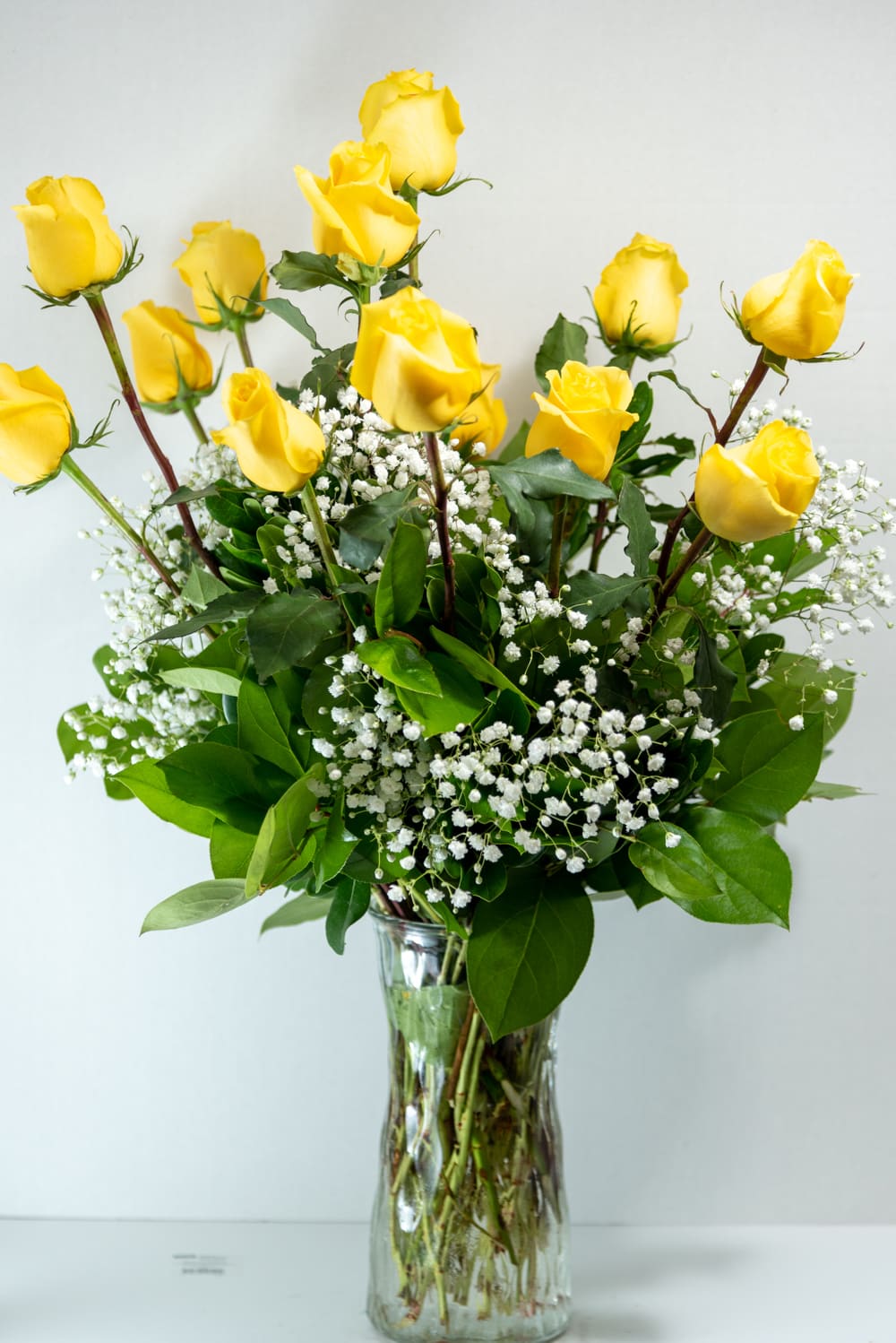 Tall arrangement of yellow roses with white filler flowers in a clear glass vase