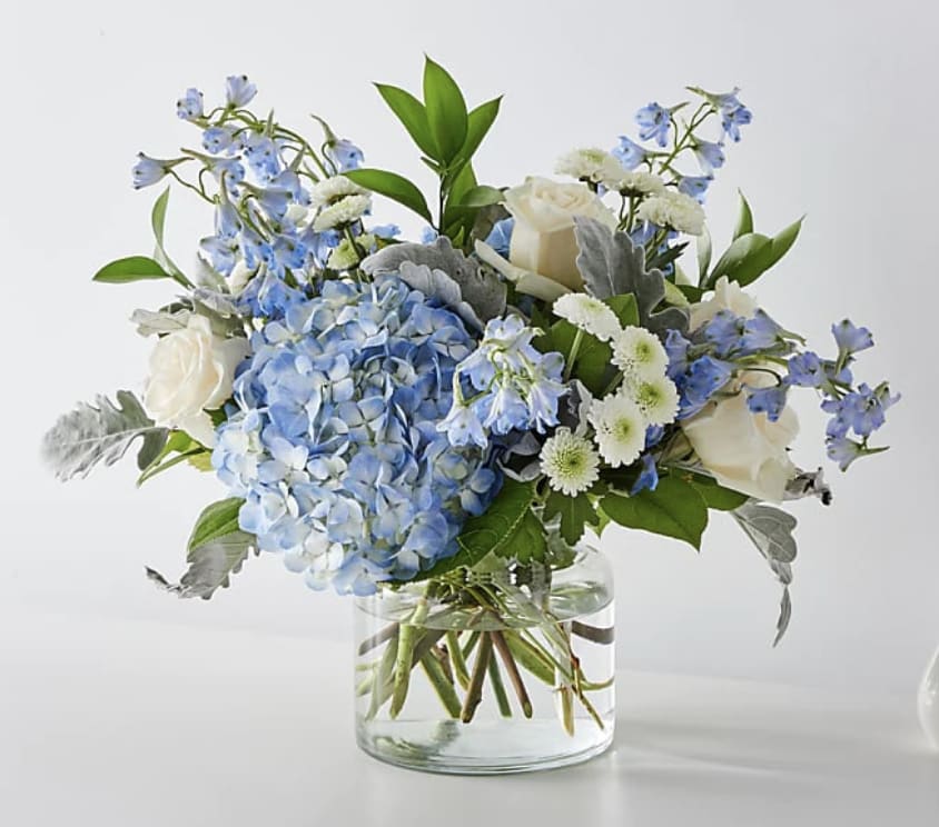 Low glass vase of blue hydrangea, white roses, white mums, and blue delphinium.