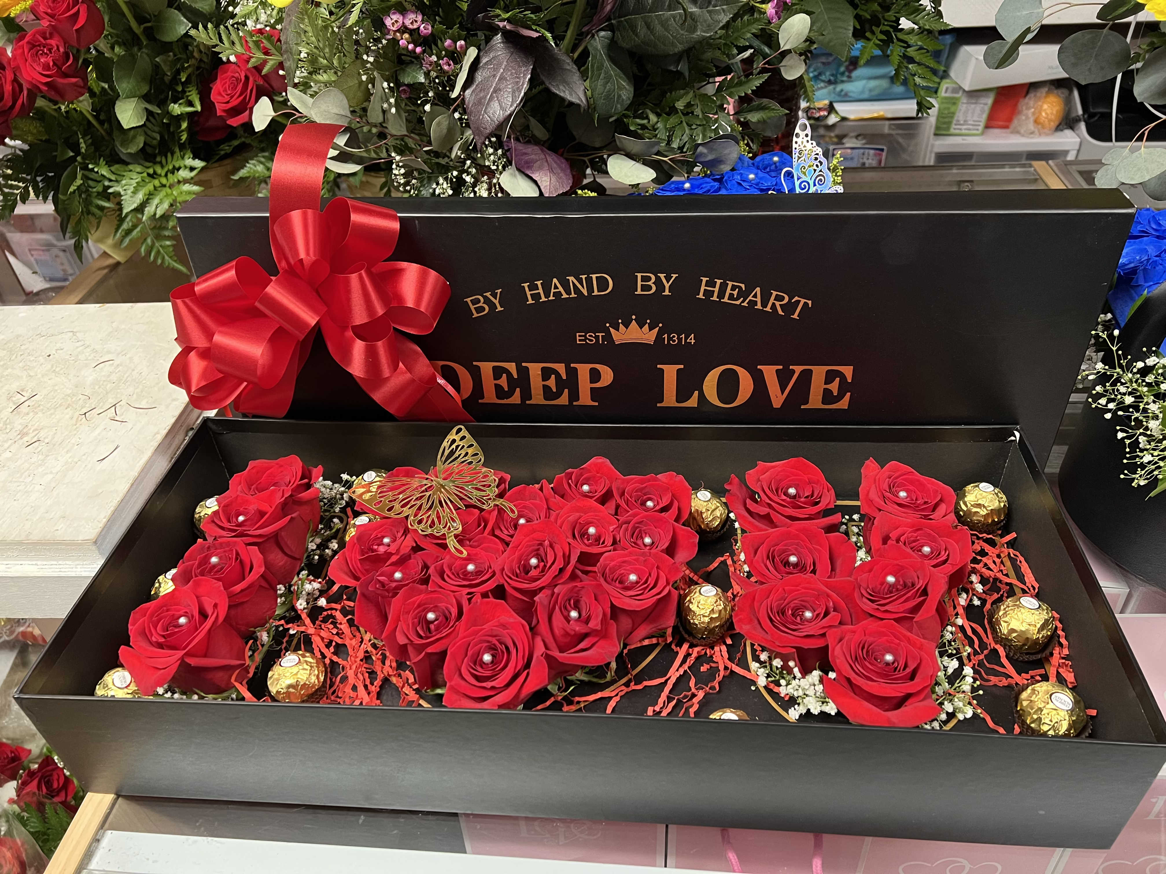I Love You Box - A low black rectangular gift box filled with lush red roses creates an immediate romantic statement-perfect as a love and romance arrangement or a Mother's Day surprise. The compact "I Love You" style presentation reads intimate and luxurious, the kind of floral gift that makes the person you're thinking of feel cherished and deeply seen for anniversaries, Valentine's Day, or any day you want to say "I love you."  Bright red roses sit snugly in a bed of red shredded paper and are dotted with small white filler blooms and pearl-like bead accents for delicate contrast. Gold-wrapped chocolates are tucked between blooms and a gold decorative butterfly pick adds a warm metallic flash, while a large red satin ribbon bow at one corner finishes the elegant look. The arrangement is presented in a low black box labeled "DEEP LOVE," offering a polished keepsake-ready display that's easy to place on a table or nightstand.  Give this to a partner, mother, or someone special who enjoys classic romantic styling-the combination of roses, chocolates, and ribbon sends a clear message of affection and celebration. The low-box format makes care simple: keep water supply as directed by the sender and avoid direct heat; ideal for same-week gifting during Mother's Day, Valentine's Day, anniversaries, or whenever you want to make someone feel treasured.
