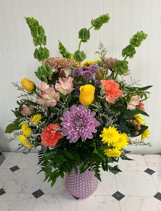 Mama's Petal Party - A joyful celebration of color and flowers! Mama’s Petal Party features a lively mix of yellow, lavender, peach, green, and orange blooms arranged in a beautiful iridescent dimple vase. Bright, fun, and full of personality, this arrangement is perfect for Mother’s Day or celebrating someone special.  Vase color may vary- pink, purple, and green.