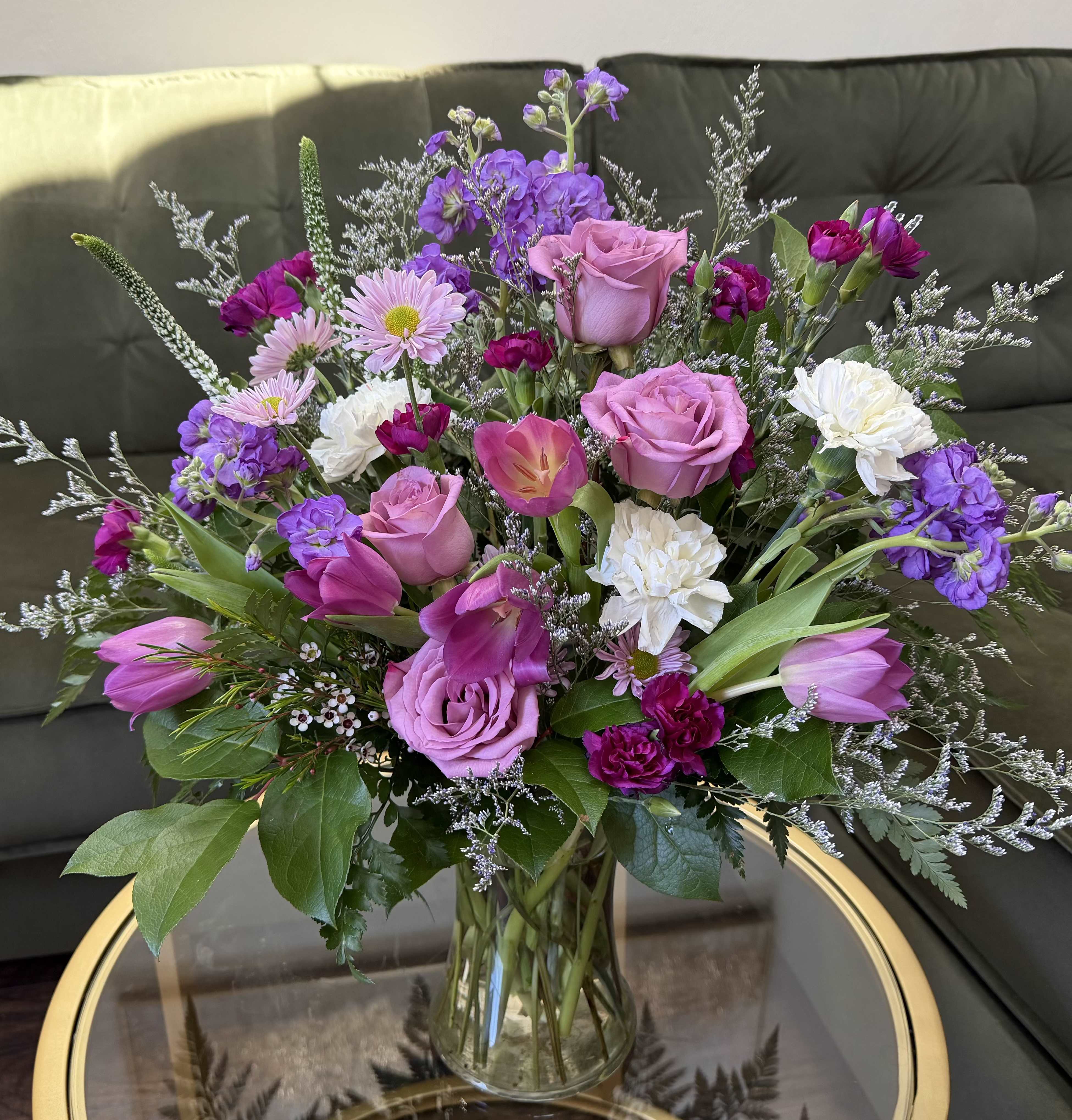 Lavender Daydream - This arrangement feels like a soft spring garden gathered at its peak—fresh, airy, and effortlessly elegant. Lavender roses take center stage, their velvety petals paired with graceful tulips in shades of rose and soft magenta, adding movement and a natural, airy flow. Delicate sprays of lavender stock and waxflower weave throughout, bringing a gentle fragrance and a touch of wild charm.  White carnations and dainty pink daisies lighten the palette, creating balance and a soft contrast, while pops of deeper berry-toned mini carnations add richness and depth. Lush greenery frames the composition, giving it that just-picked, organic feel.  Designed in a clear glass vase, this piece has a loose, garden-inspired silhouette—refined yet relaxed. It’s the kind of arrangement that feels warm, feminine, and quietly luxurious, perfect for celebrating meaningful moments, as Mother's Day and Birthday gifts or simply elevating everyday spaces with beauty.
