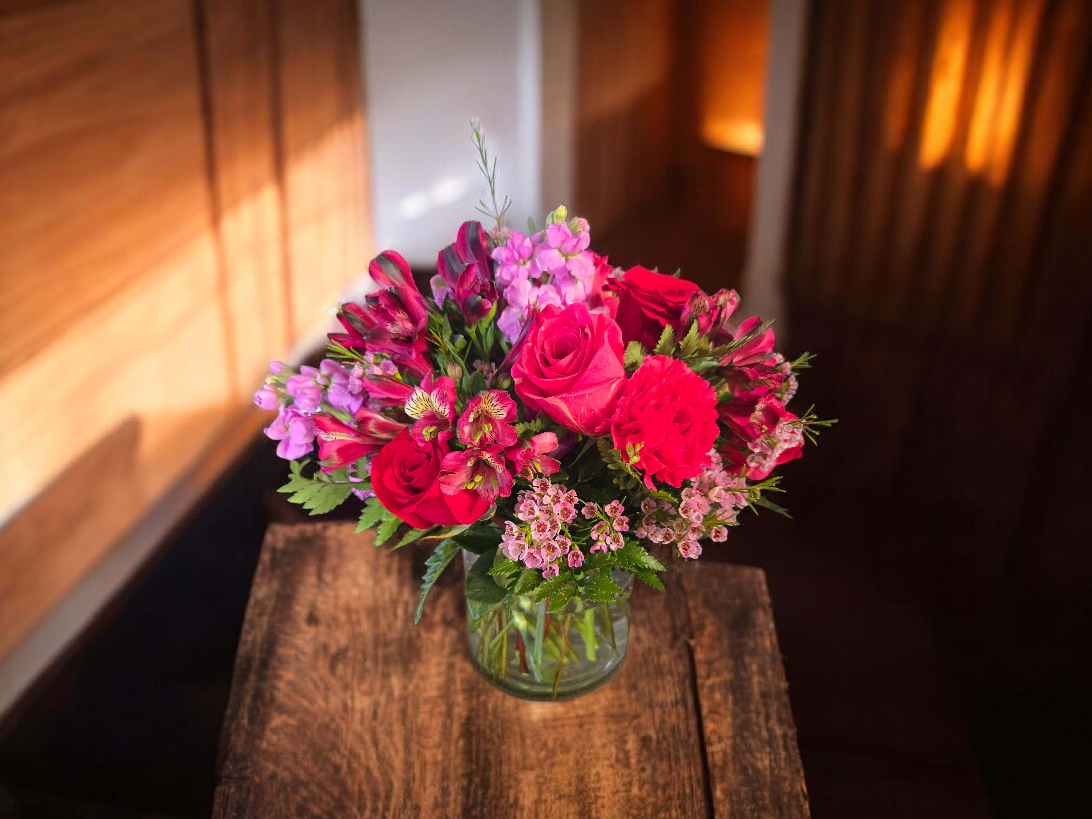 Blushing Romance Bouquet - A lush, rounded bouquet of pink, red, and purple blooms that reads like a blushing love letter-perfect as an anniversary roses arrangement, a birthday surprise, or a just‑because gift to celebrate romance. The clear glass vase keeps the look polished and lets the vivid colors take center stage for Mother's Day, Women's Day, or any moment you want to make her feel truly seen.  Roses anchor the center while clustered stock, alstroemeria, and carnations weave texture and striped detail for a layered, garden‑like feel. Compact and dense, this arrangement arrives ready to display in the included clear vase; light daily watering and fresh water changes help keep blooms vibrant for days.