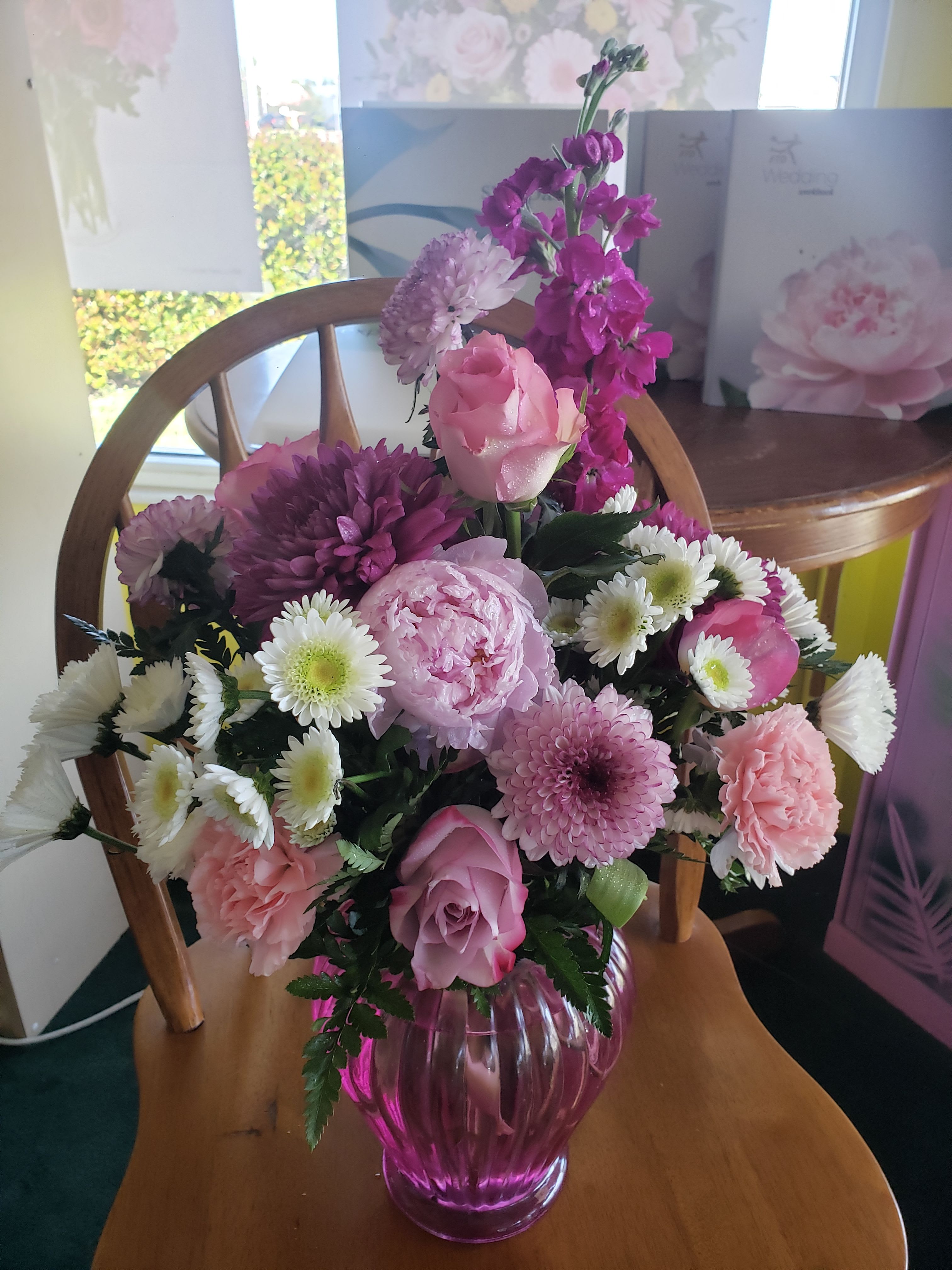 BEAUTY LAVENDER BOUQUET - A lush cluster of pink, lavender, purple, and white roses, carnations, chrysanthemums, and daisy-like blooms greets the eye-perfect as a birthday bouquet or anniversary flowers to make someone feel genuinely celebrated. The dense, rounded composition reads romantic and cheerful: soft petals and airy accents mix to create a feminine, garden-fresh impression that says "thinking of you" with warmth and charm.  This arrangement arrives gathered in a ribbed pink/purple glass vase that adds subtle shine and a vintage-modern touch to tabletops or mantels. Large focal roses sit alongside textured carnations and chrysanthemum blooms, with small daisy-style flowers providing light, playful contrast and brighter magenta accents for energy. The design is tightly clustered and abundant, giving a classic florist-style look that's ready to display-no extra arranging needed.  Ideal for birthdays, anniversaries, bridal showers, or simply brightening a loved one's day, this bouquet suits recipients who appreciate soft, romantic color and full, touchable texture. Care is straightforward: keep stems trimmed and vase water fresh to extend the life of the mixed blooms and enjoy their layered colors for days.