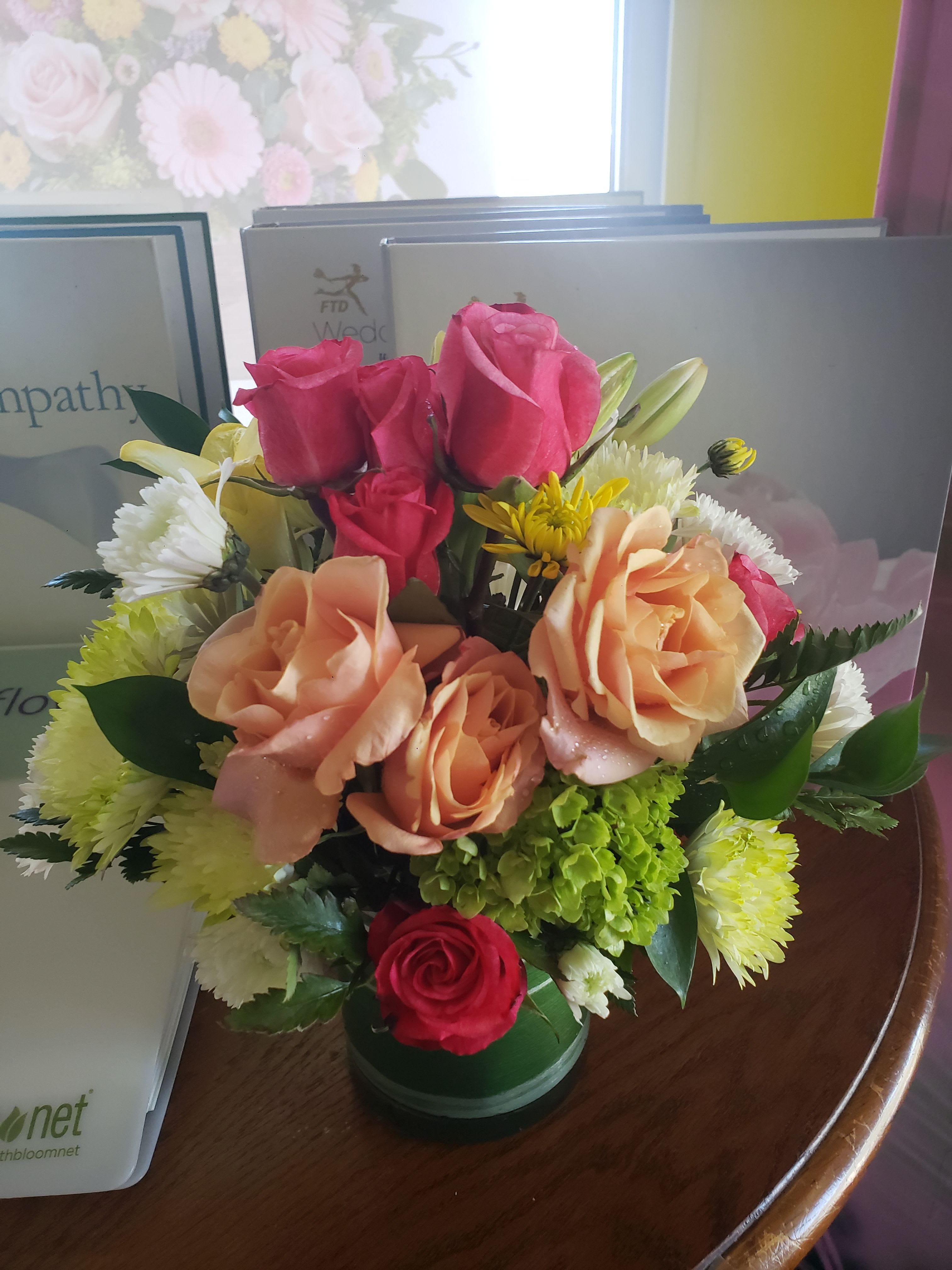 BEAUTY ORANGE AND GREEN BOUQUET - Hot pink and peach roses take center stage in this rounded sympathy bouquet, a warm yet respectful flower arrangement designed for condolence or remembrance. The soft white and sunny yellow chrysanthemums-layered with a hint of green hydrangea near the center-give the piece a tender, uplifting presence that reads as both comforting and gently celebratory.  Arranged tightly in a low glass vase (shown also in a green vase option), the compact shape creates a full, abundant look that feels polished without being fussy. Lilies and mixed textures add fragrance and depth while the included sympathy card lets you send a thoughtful message; colors span hot pink, peach, white, and yellow for a balanced, hopeful palette.  This bouquet suits memorial services, bedside visits, or as a quiet way to say you're thinking of someone-yet its cheerful tones also make it appropriate for birthdays, anniversaries, or springtime gestures. Available in the pictured low vase size; please follow the care instructions on the enclosed card to keep blooms fresh for as long as possible.