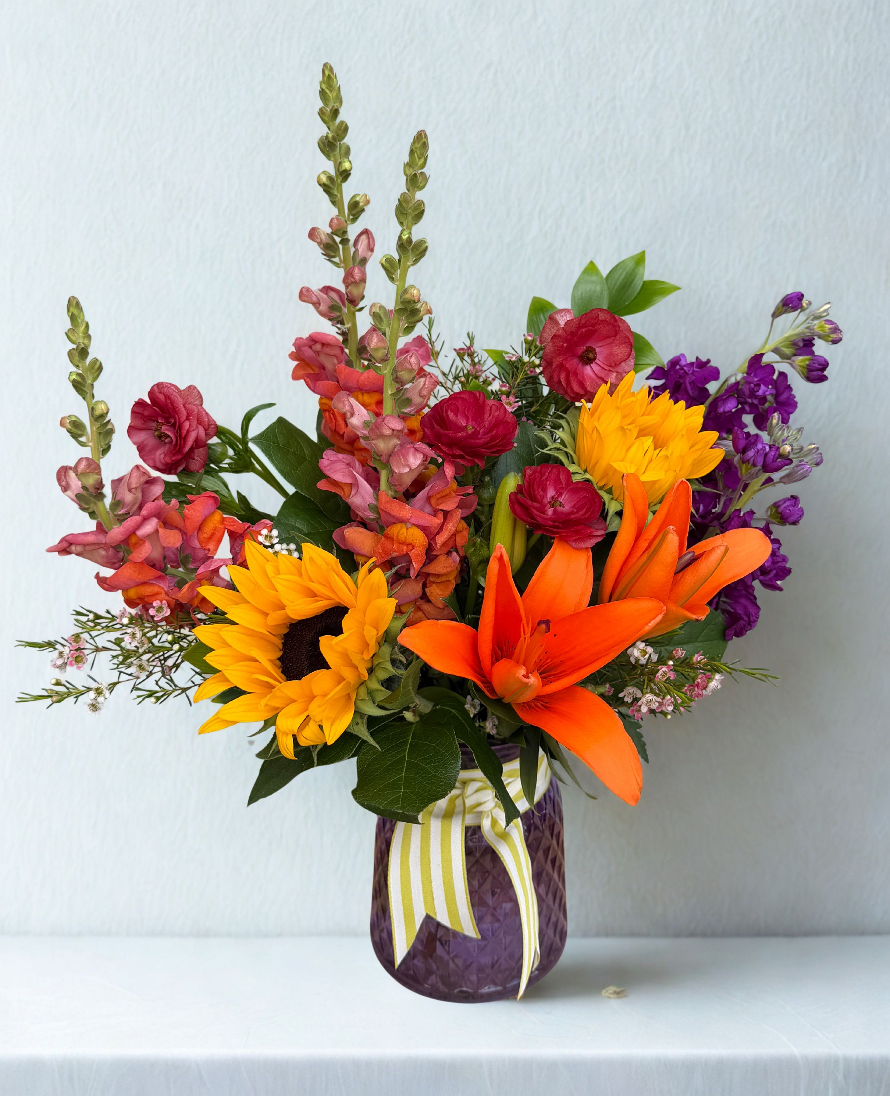 Hillsides Happy Blooms - A vibrant celebration of color, this eye-catching arrangement features bold orange lilies, sunny sunflowers, and rich jewel-toned blooms accented with lush greenery. Designed in a textured vase and finished with a cheerful ribbon, it brings warmth, energy, and a touch of whimsy to any space. Vase color may vary