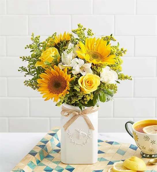 Yellow sunflowers and roses arranged in a white vase with a ribbon