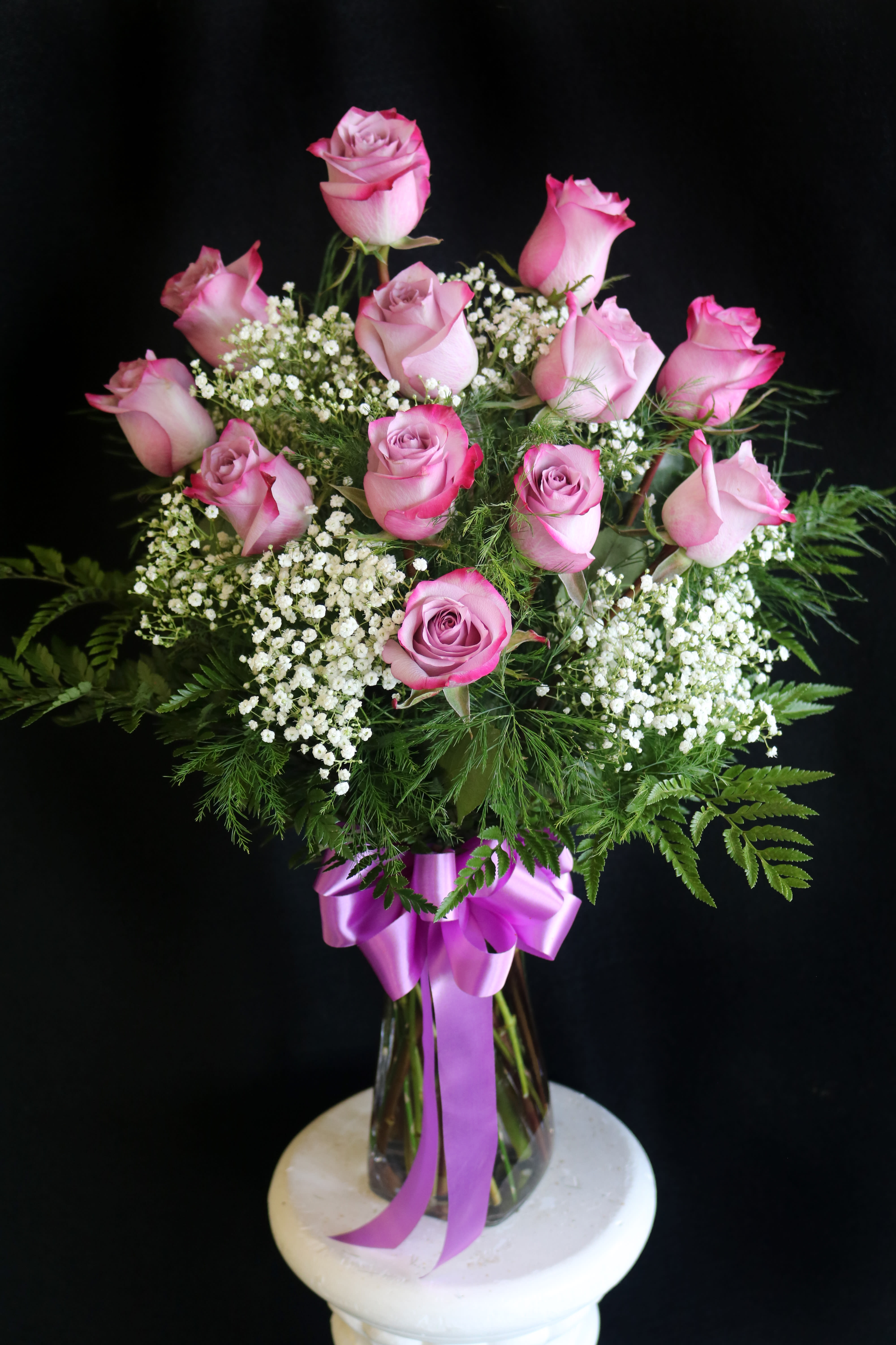 Pink roses in a glass vase with white filler flowers and a purple ribbon