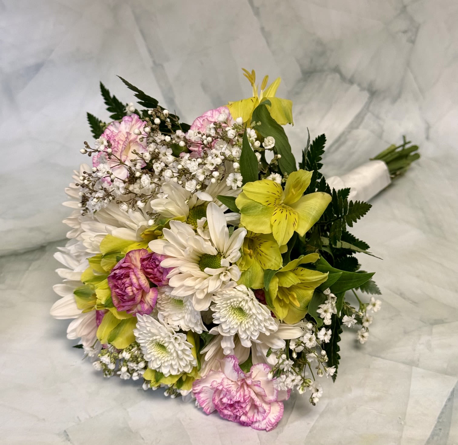 Spring Colors Prom Bouquet - A bright, hand-tied prom bouquet that feels like a pocketful of spring - ideal as a prom corsage alternative or a cheerful gift for birthdays, thank-yous, and spring celebrations. The focal mix of white daisy-like blooms and vivid yellow alstroemeria gives the arrangement a sunny lift, while soft pink carnations add a touch of gentle romance the recipient will notice right away.  Packed into a full, rounded shape, this arrangement pairs large white daisies with smaller pink carnations, bright yellow alstroemeria, and airy clusters of tiny white filler flowers for texture and movement. The palette of white, yellow, pink, and green reads fresh and lively; stems are neatly finished with a white bouquet wrap for an easy, hand-held presentation that arrives ready to give.  Perfect for the person you want to make feel celebrated - whether it's prom night, a birthday, or a simple "thinking of you" moment - the Spring Colors Prom Bouquet offers a fuss-free way to brighten someone's day. Treat gently, recut stems and place in fresh water on arrival to keep blooms looking their best.
