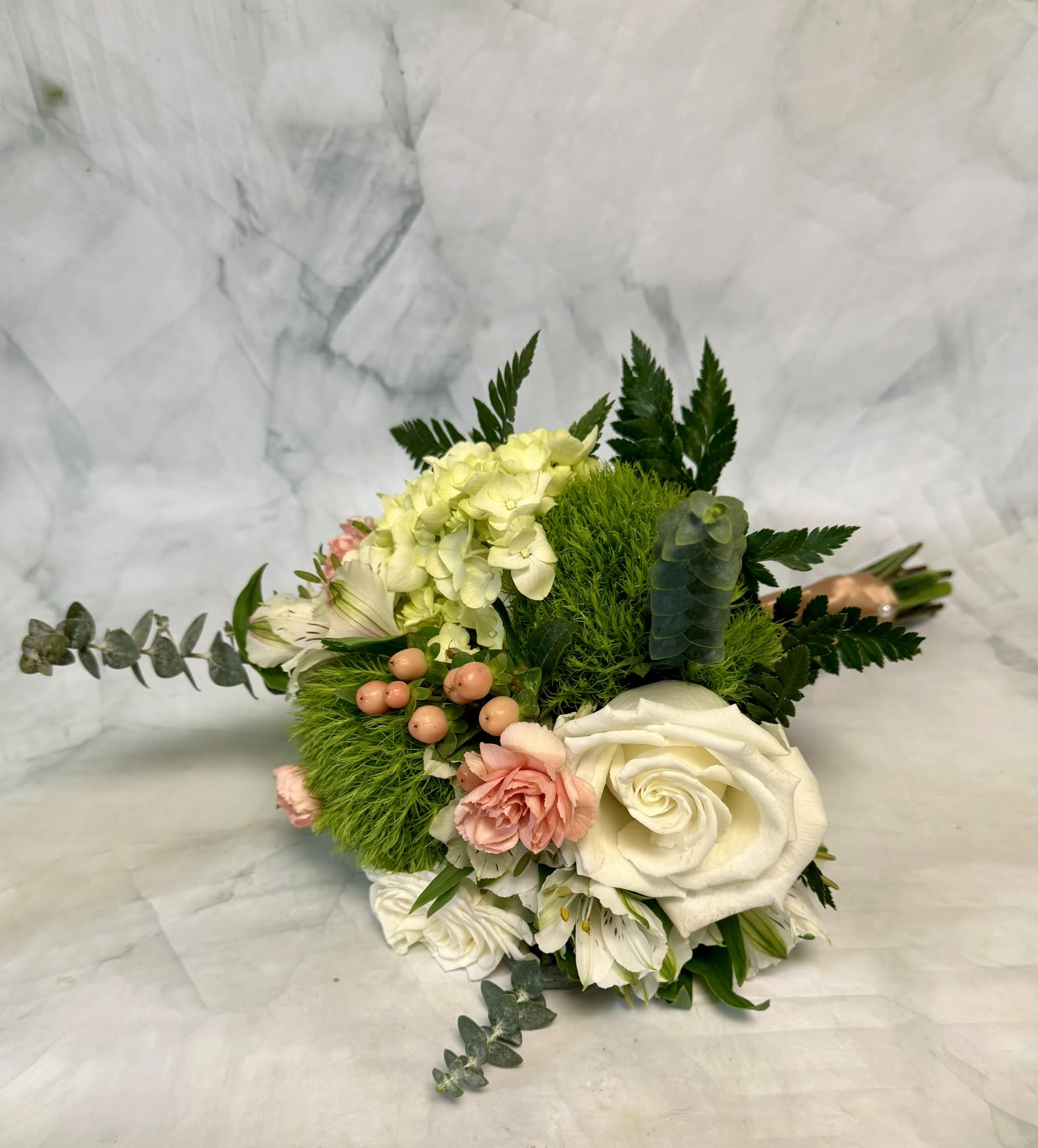 White, Peach and Green Prom Bouquet - A lush, garden-style prom bouquet that reads as quietly romantic-perfect for prom, a wedding, or an anniversary moment when you want the recipient to feel seen and softly celebrated. This hand-tied bouquet places creamy white roses and pale hydrangea at its center, with blush carnations and white alstroemeria layered around them to create a compact, rounded silhouette that looks elegant in photos and natural in person.  Textural contrasts give the bouquet personality: bright mossy-green filler and dark fern-like foliage frame the soft cream and pink blooms, while small peach berry accents add subtle color punctuations. The palette of white, peach, blush, and fresh green keeps the feeling light and garden-fresh. Arranged low and full in a ceramic vase, it reads intimate and lush-an ideal choice for someone who prefers romantic understatement over flash.  Give this to the person you want to make feel cherished-whether it's for prom photos, a vow renewal, or a gentle sympathy expression-and they'll notice the thoughtful layering and soft scent. The compact hand-tied design sits beautifully in the included ceramic vase; treat stems with fresh water and trim occasionally to keep blooms looking their best.
