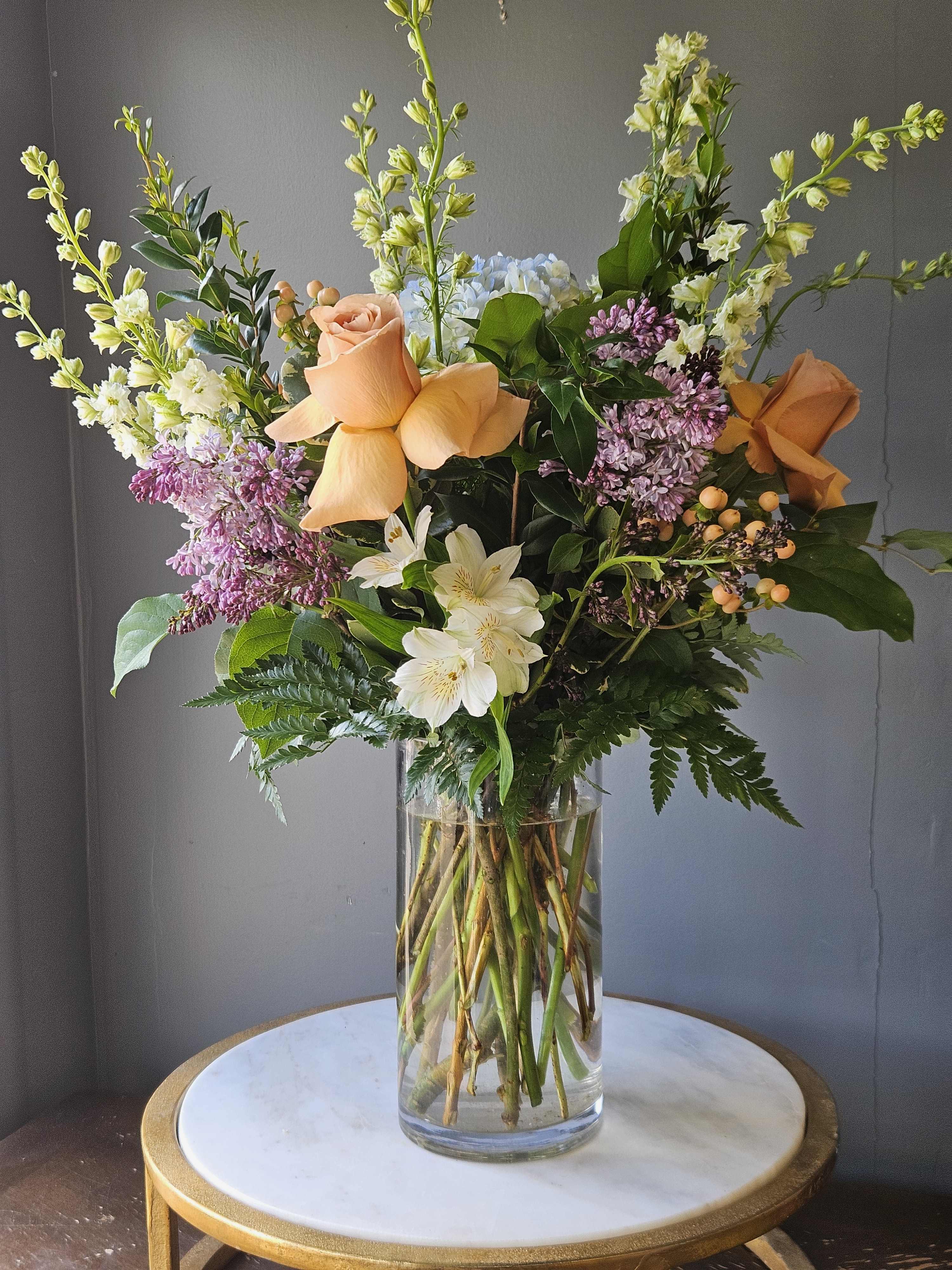 Tall mixed bouquet with peach roses, white blooms, lilac stems in a clear glass cylinder vase.