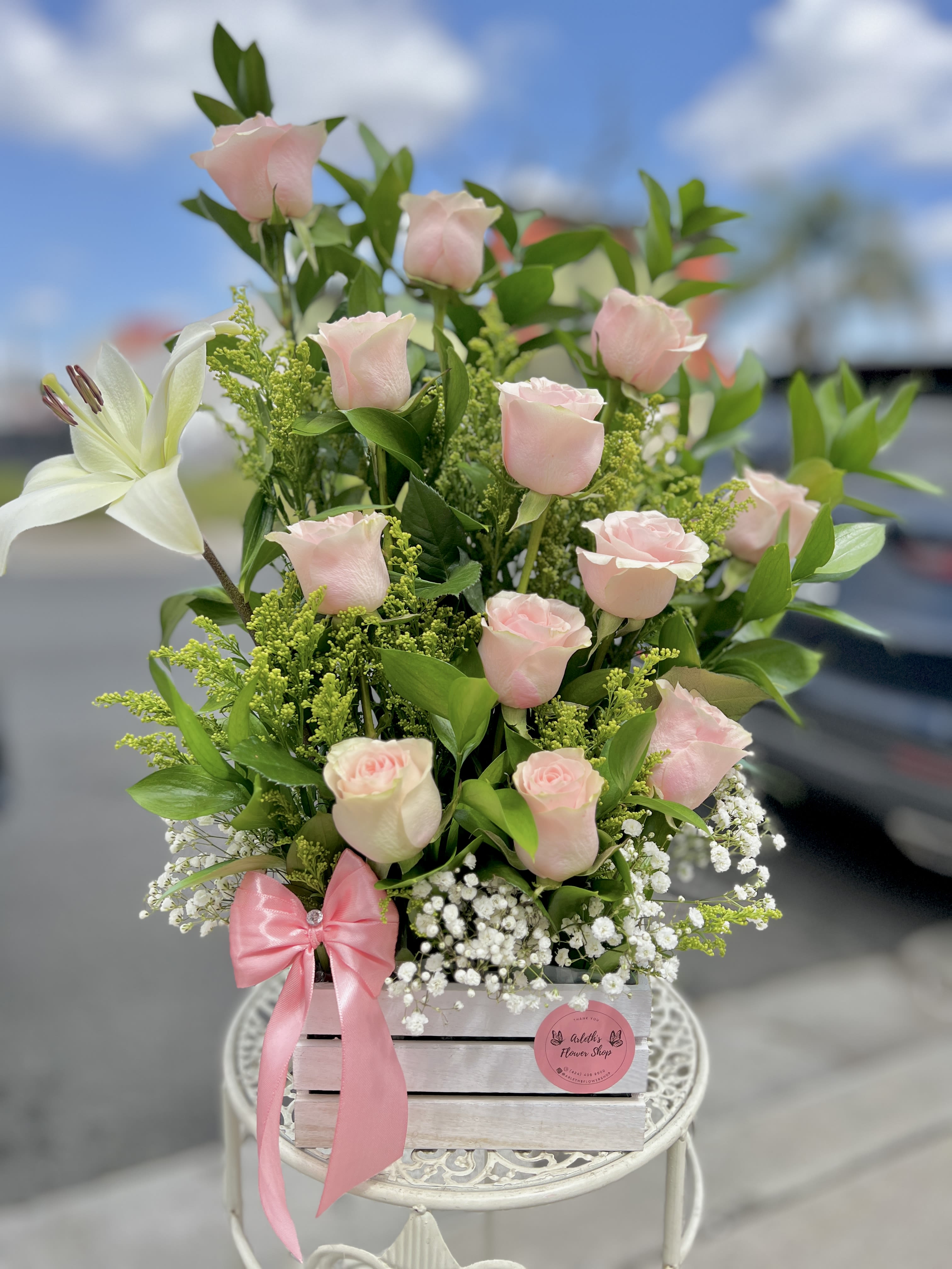 Pink roses and a white lily arranged in a wooden box with a pink ribbon.