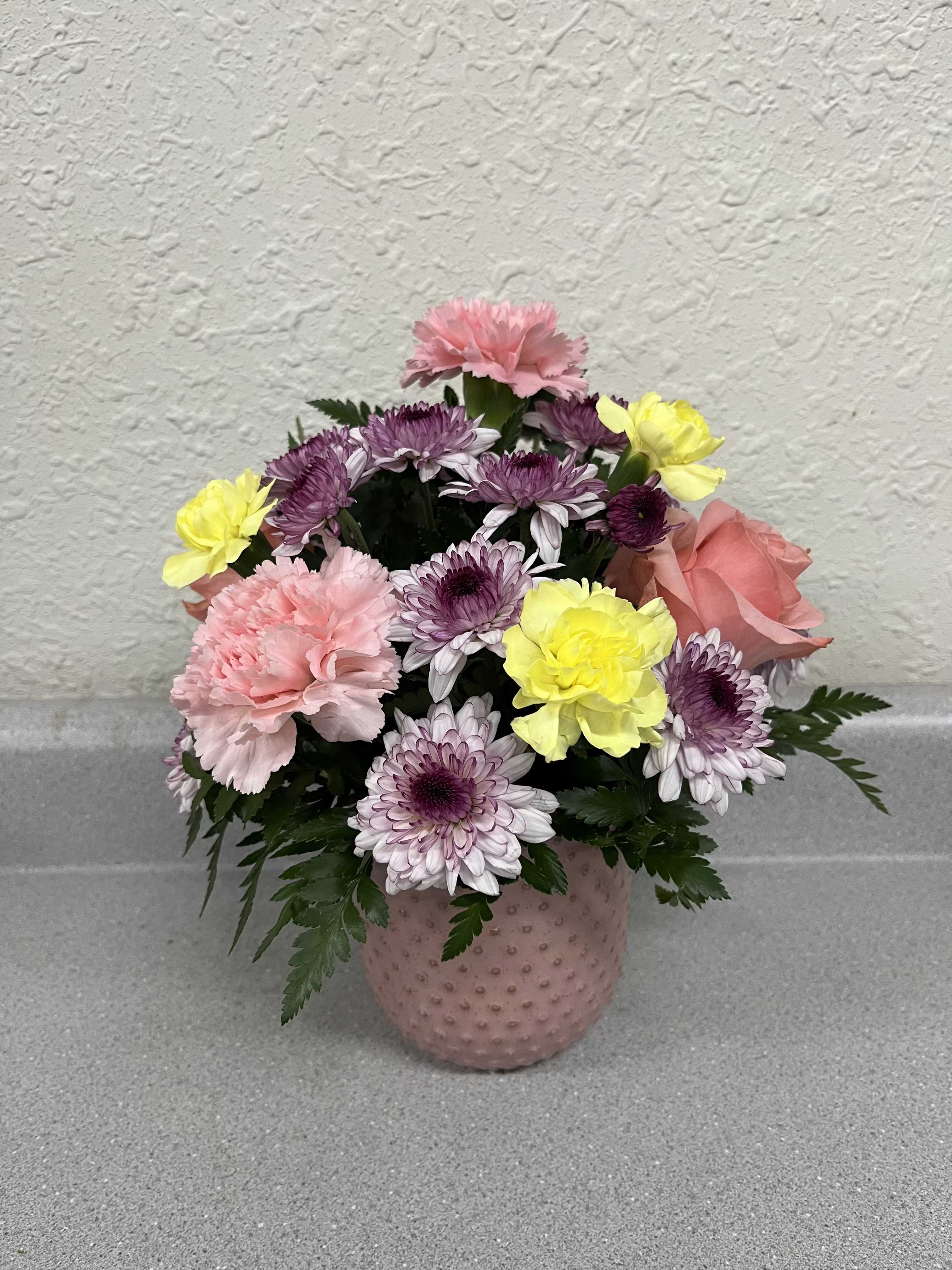 Petals Of Love - This cheerful dome of pastel blooms includes pink carnations, lavender-and-white chrysanthemums, sunny yellow accents, and a soft pink rose making this flower arrangement a perfect bouquet for any occasion including Mother's Day. The rounded pink textured ceramic vase gives the design a polished, elegant look that shows your love and appreciation.  