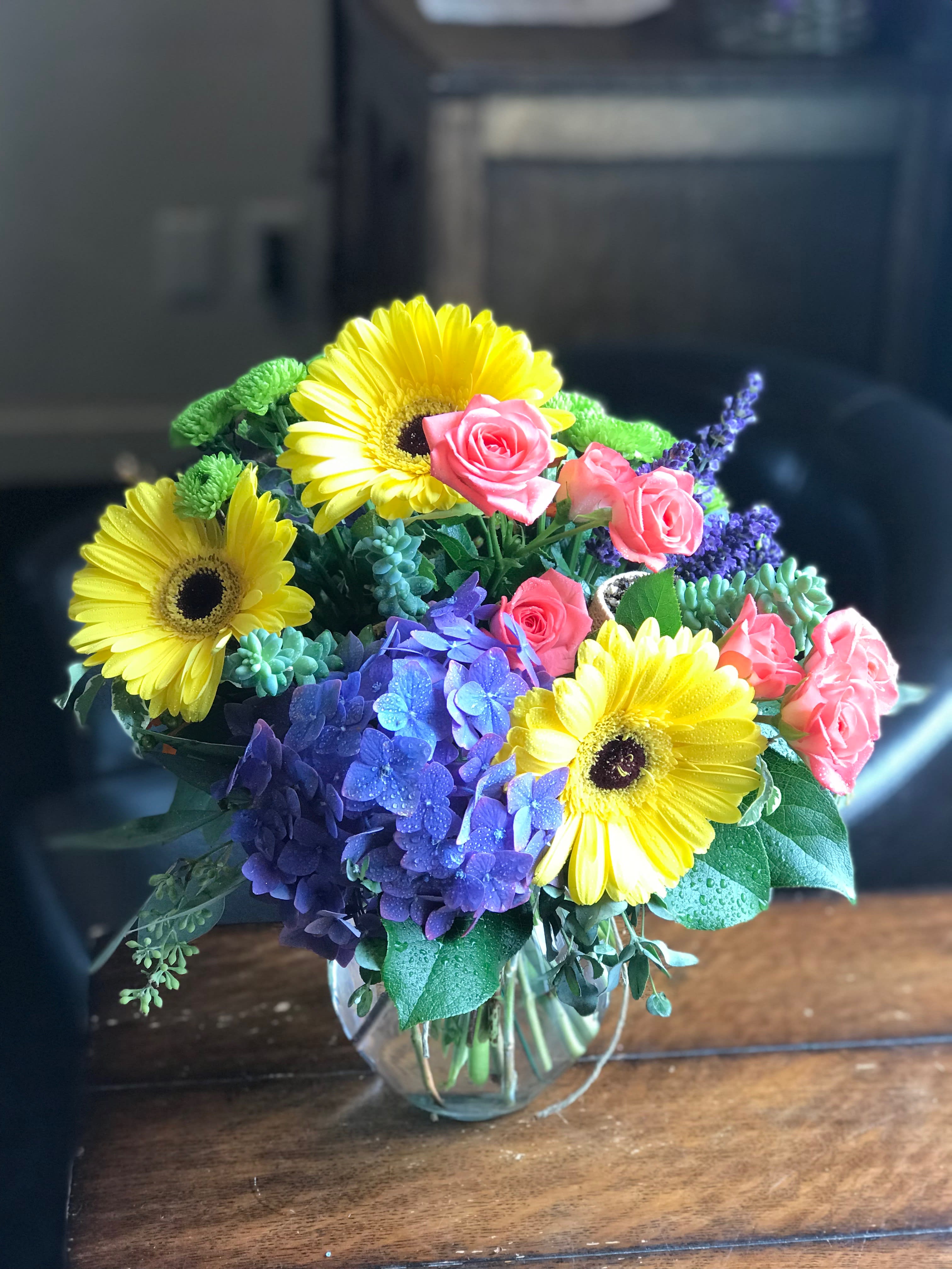 Colorful bouquet of yellow gerbera daisies, pink roses, and purple hydrangeas in a glass vase