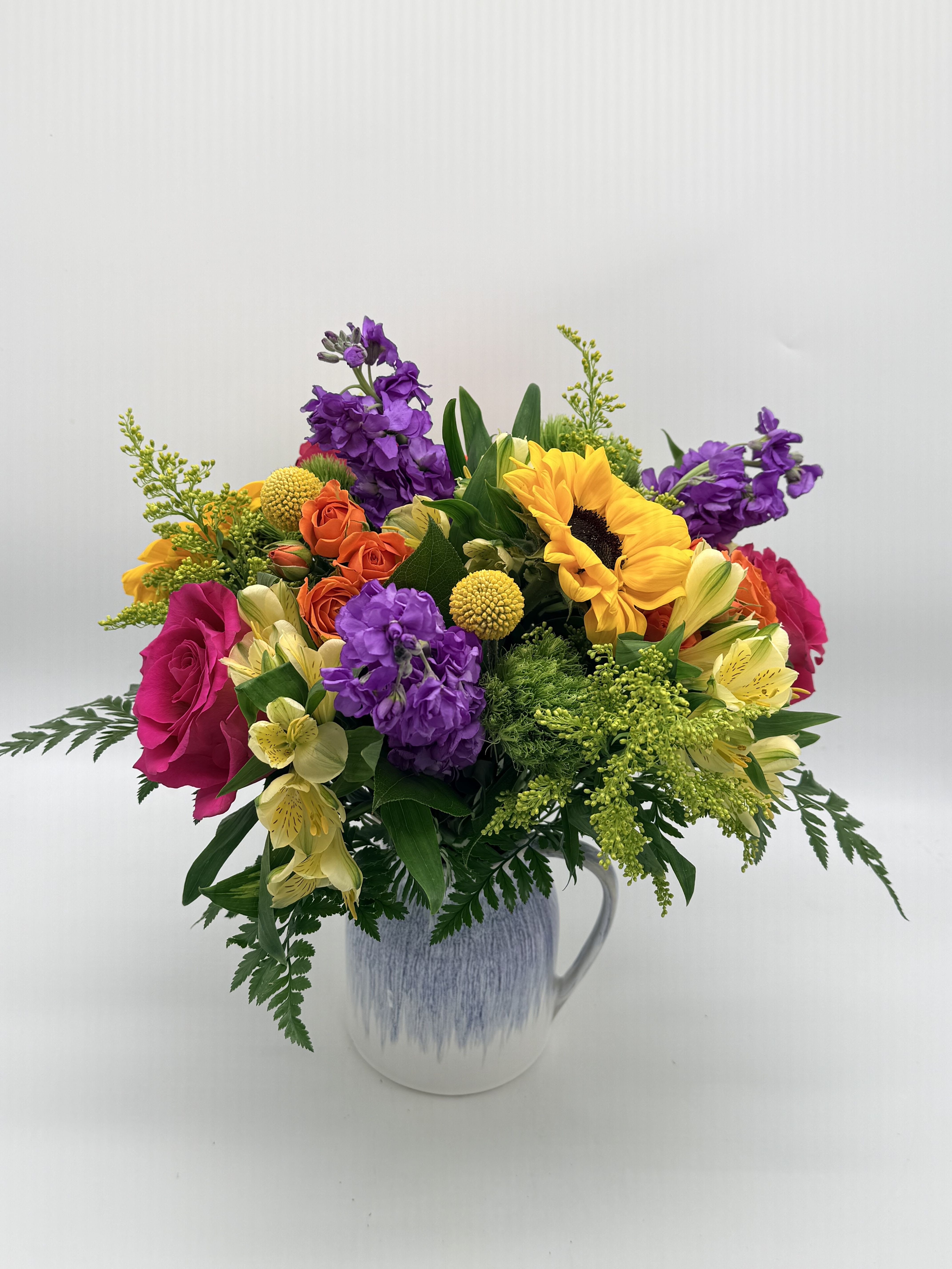 Joyful Pitcher - Bright, cheerful, and full of charm, this vibrant arrangement is beautifully designed in a keepsake ceramic pitcher for a fresh, garden-inspired look. Featuring a stunning mix of hot pink roses, golden sunflowers, vivid orange spray roses, and rich purple blooms, this design overflows with color and joyful energy. Accented with textured greenery and playful filler, it feels like a hand-gathered bouquet straight from a sunny garden. Ideal for birthdays, get well wishes, celebrations, or just because, this arrangement is sure to brighten any space and lift any mood.  Flower varieties and colors may vary slightly based on seasonal availability while maintaining the overall style and vibrant palette.