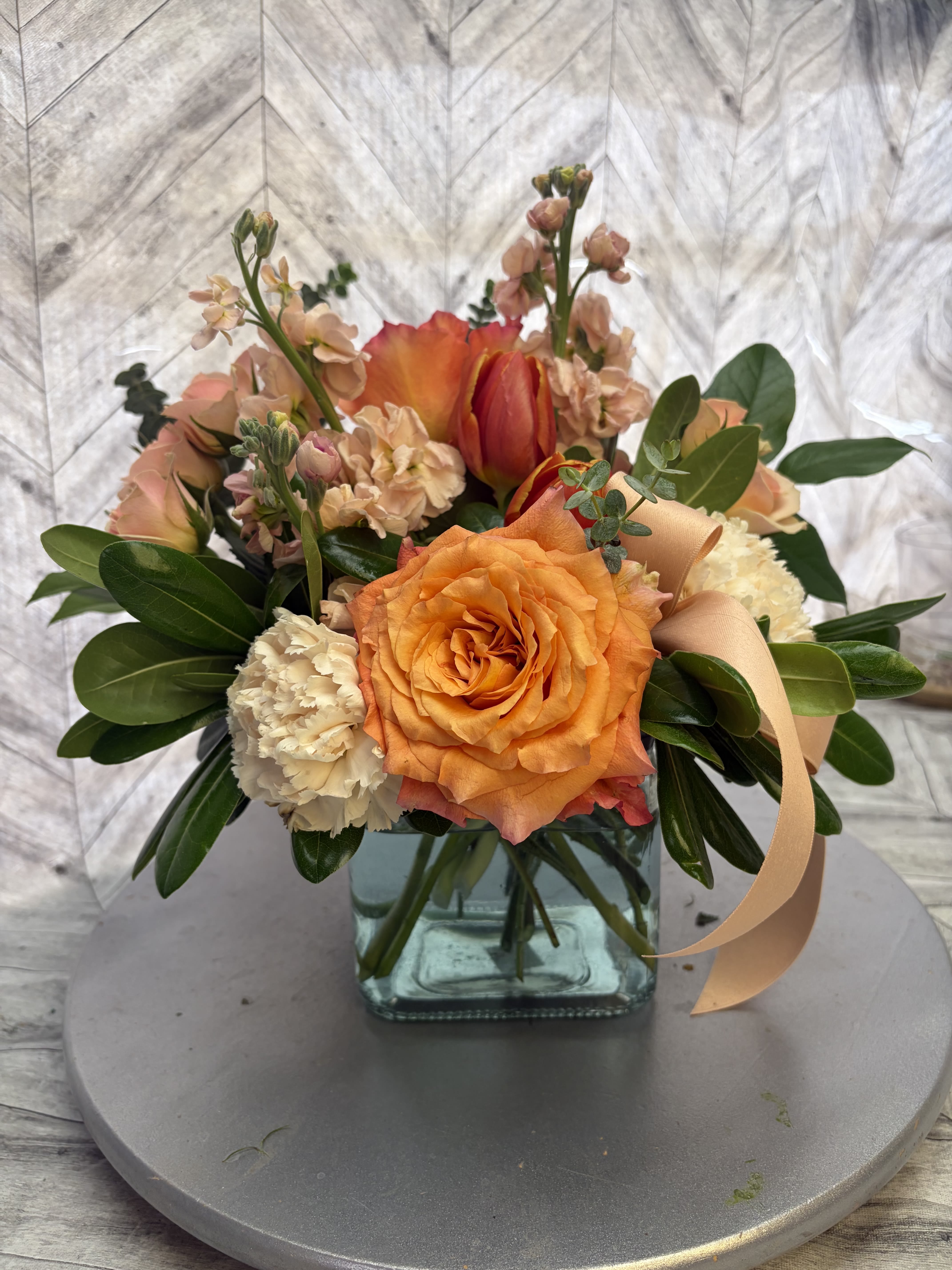 Donna's Delight - This stunning handcrafted bouquet captures the warm glow of a sunset with its rich blend of orange, peach, and cream tones. At the heart is a beautiful large orange rose, surrounded by soft peach stock flowers, creamy white carnations, delicate smaller blooms, and lush green foliage for texture and contrast. A flowing peach satin ribbon adds a touch of elegance and softness.  