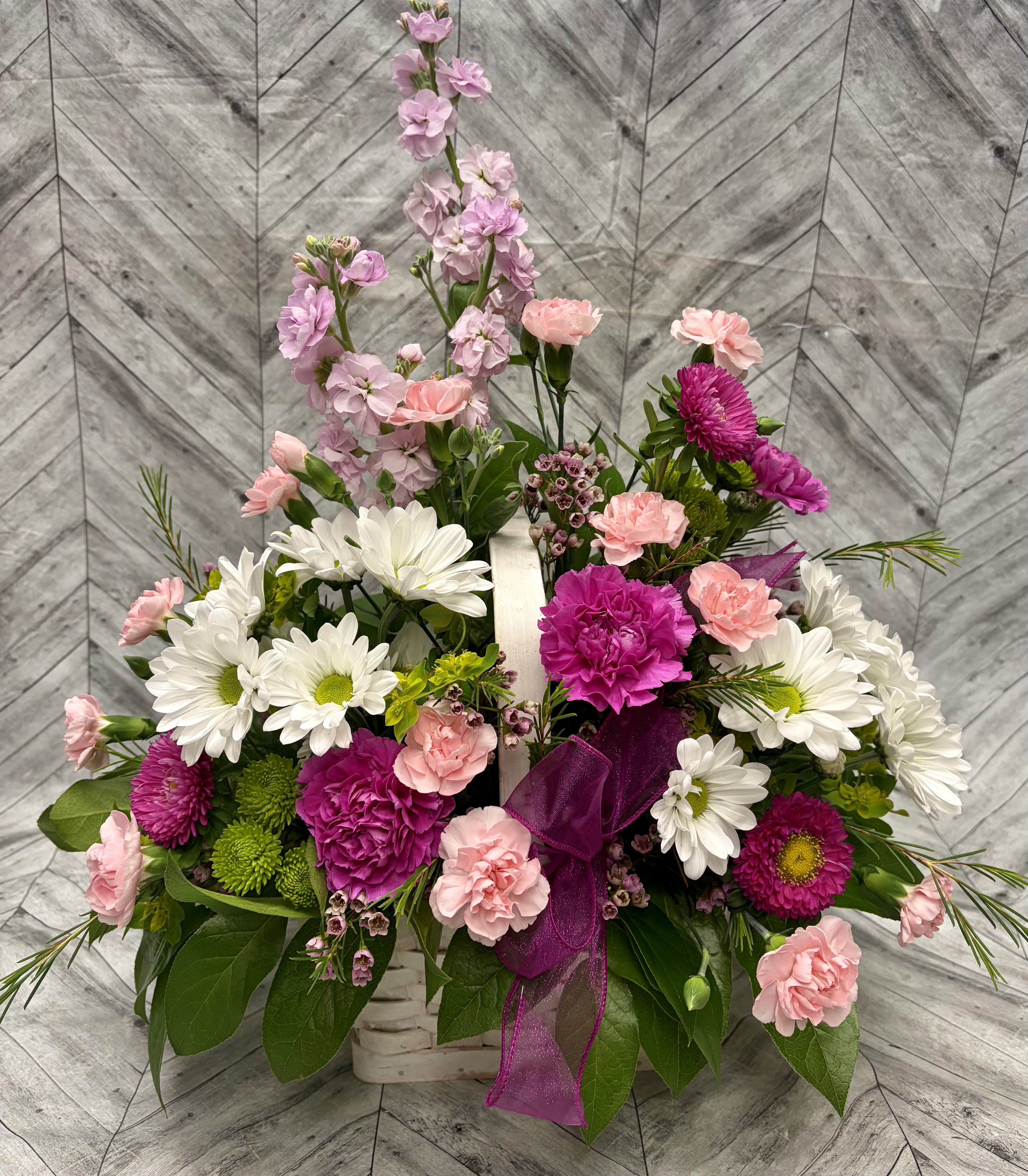 Donna's Basket of Joy - Celebrate the special woman in your life with this overflowing basket of fresh flowers bursting with joy and color!  This stunning arrangement features a harmonious blend of vibrant magenta carnations, soft pink roses and stock, crisp white daisies, delicate purple accents, and lush green foliage. Nestled in a charming white wicker basket and finished with a luxurious sheer purple ribbon bow, it creates a warm, feminine, and heartfelt gift that speaks volumes of love and appreciation.  