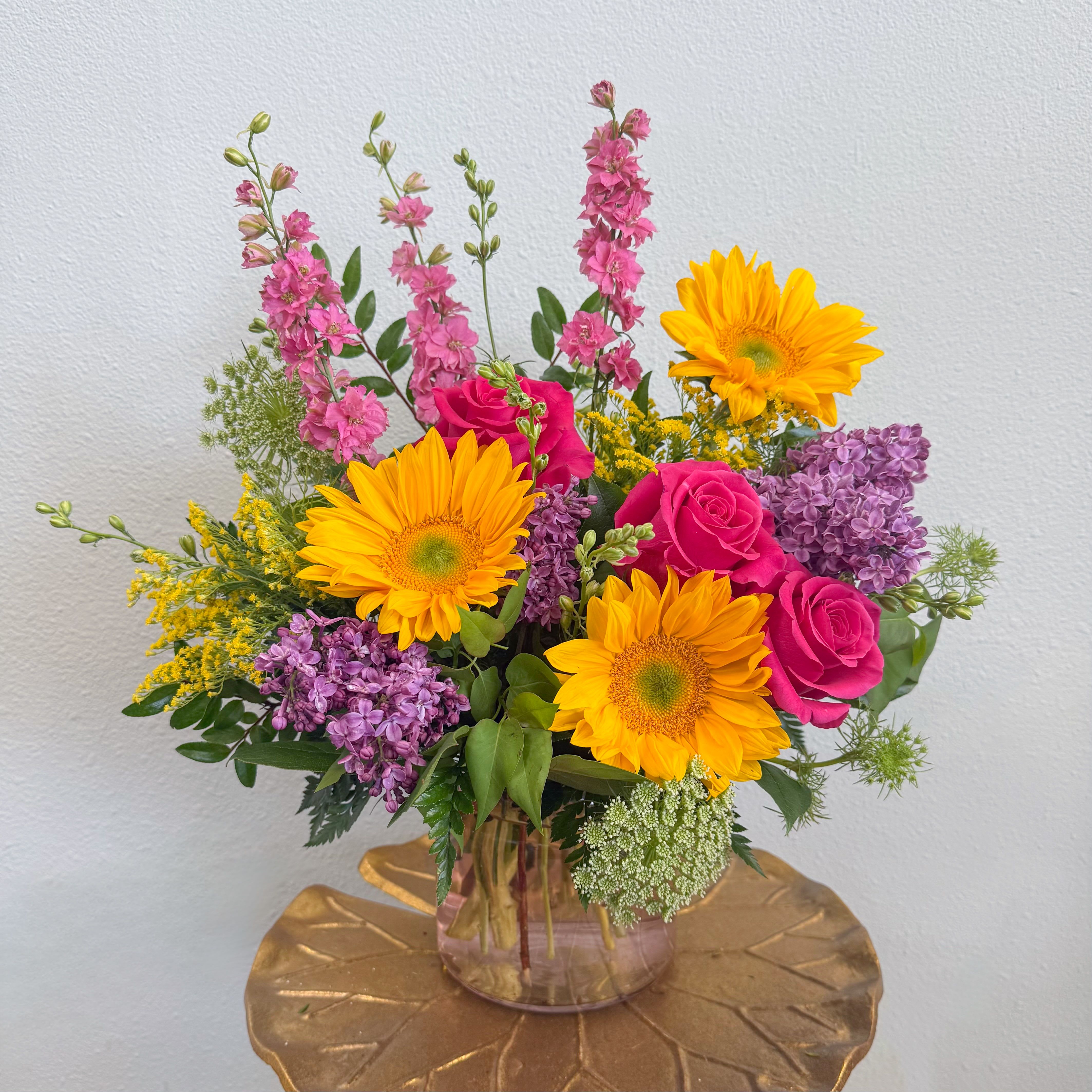 Golden Hour - A radiant, sunlit arrangement inspired by the warmth of late afternoon light. Golden sunflowers and soft garden textures are layered with roses and seasonal blooms, creating a design that feels both joyful and effortlessly refined.  Bright yet balanced, Golden Hour brings a natural glow to any space. 