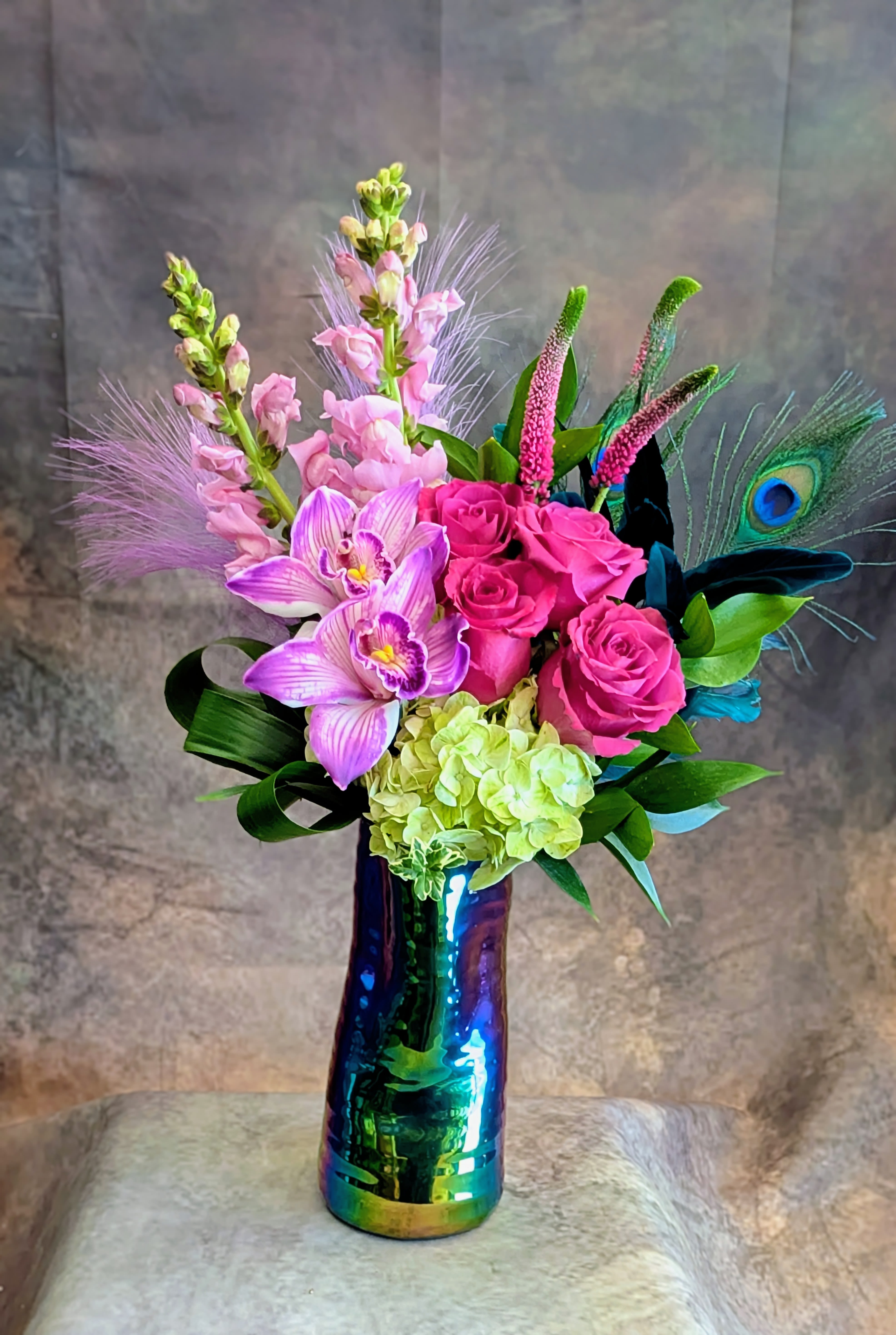Zara - Bold, vibrant, and undeniably modern, Zara is a striking floral arrangement designed in rich jewel tones with an artful, exotic edge. Featuring premium Blueberry roses in deep plum hues, this design is accented with elegant purple cymbidium orchids, soft green hydrangea, and delicate snapdragons for a lush, dimensional composition. Textural touches of veronica, tropical greenery, and a dramatic peacock feather add movement, height, and a distinctive, high-end finish. Arranged in an artistic iridescent ceramic vase, Zara is perfect for birthdays, celebrations, or anyone who loves a bold, statement floral with a unique twist. Its vivid palette and sculptural design make it a standout choice for luxury flower delivery in Las Vegas. Handcrafted in Las Vegas by Windmill Floral Expressions, a locally owned and family-operated florist established in 1991, each arrangement is custom-made with the freshest flowers available—no premade designs, always one-of-a-kind.  Same-day flower delivery available in Las Vegas and Henderson, including Anthem, MacDonald Ranch, and surrounding areas.  Curbside pickup available—please call ahead to place your custom order. Perfect for: birthday flowers, modern floral arrangements, exotic-inspired designs, and luxury flower delivery in Las Vegas.