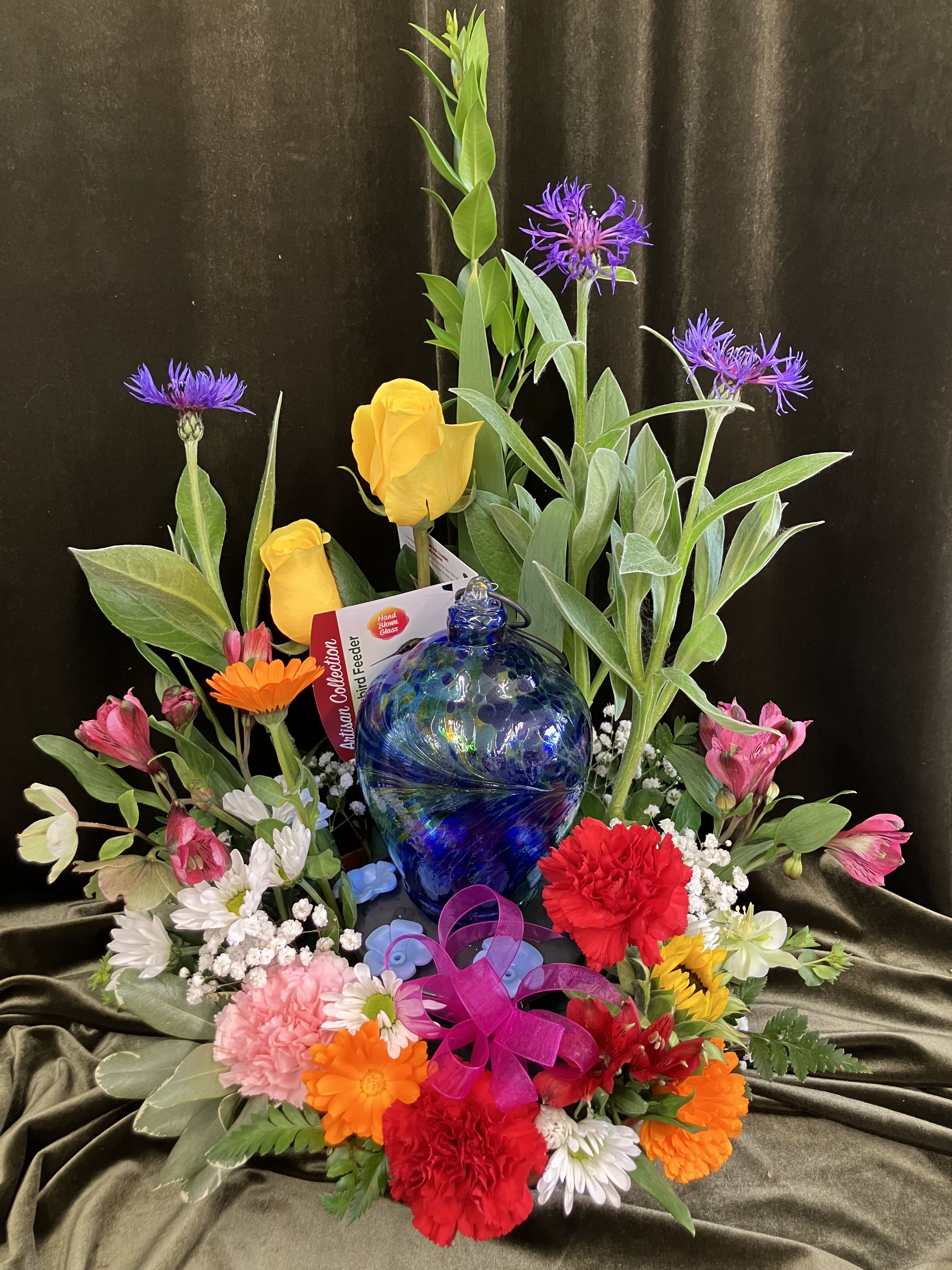 Flower Arrangement with Hummingbird feeder - We have the most wonderful arrangement for you to send this Mother's Day! Bright, fragrant, beautiful bloom surround a blue glass hummingbird feeder. Your flowers are a mixture of variety and color and will be hand picked out by the designer!
