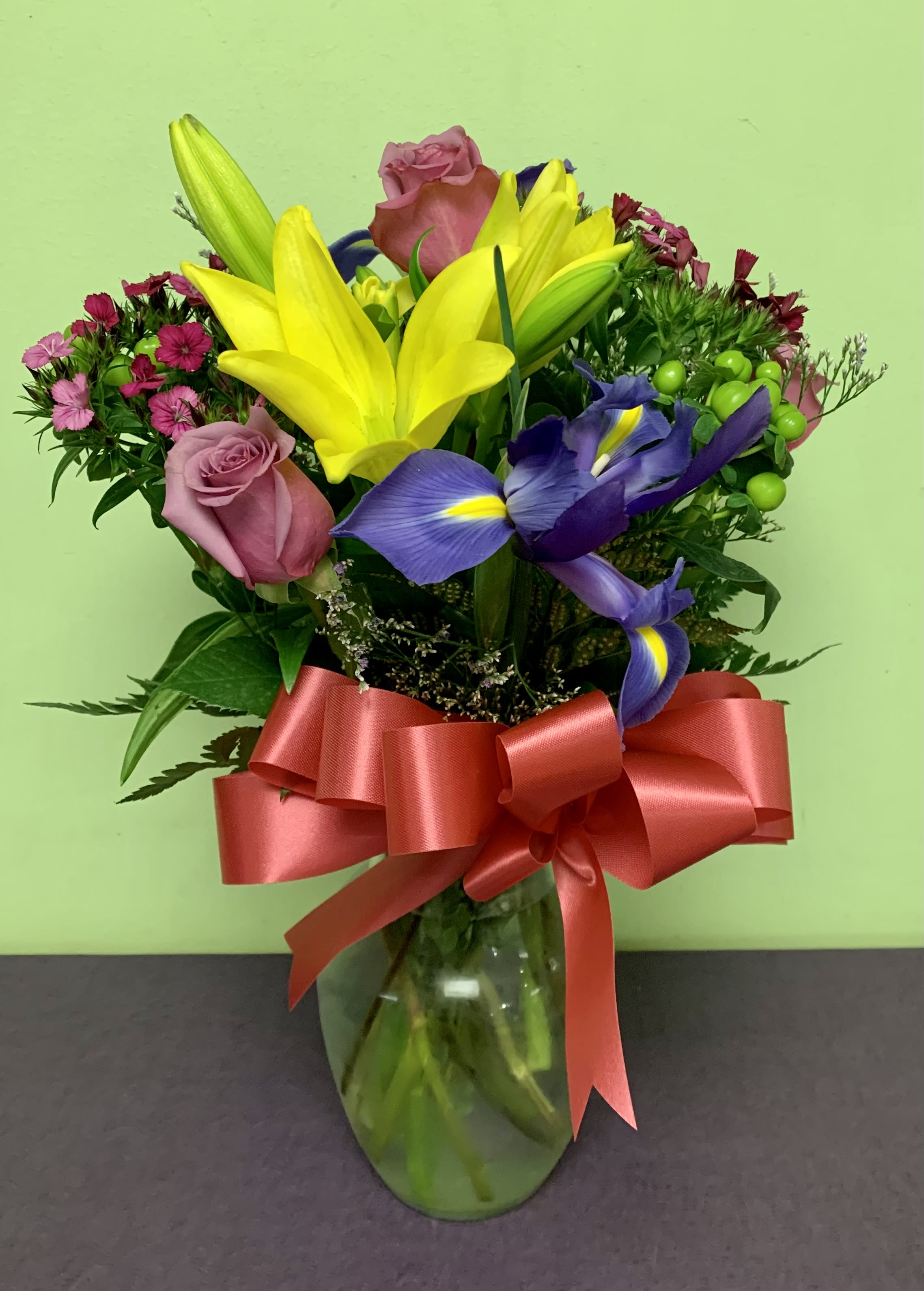 Bouquet of yellow lilies, purple irises, and pink roses in a glass vase with a red ribbon