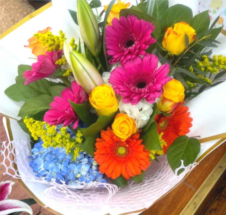 Spring Mix Bouquet - Bright, saturated color greets the recipient with this Spring Mix bouquet - a cheerful mixed-flower arrangement perfect as a birthday bouquet, congratulations gift, or bright everyday surprise. Magenta and pink gerbera daisies pair with sunny yellow roses and bold orange gerberas while a cloud of blue hydrangea anchors the composition; unopened lily buds rise through the center to add height and an elegant, structured silhouette that feels celebratory and energetic.  The hand-tied design is finished in white paper with gold trim plus pink and decorative mesh wraps for a polished, gift-ready presentation. Yellow filler blooms and broad dark-green foliage add fullness and contrast, creating a lively texture and rich color depth. Sized to feel generous without overwhelming, it's the kind of arrangement that makes the person you're thinking of feel genuinely seen and celebrated - order with standard care: keep in fresh water, trim stems, and avoid direct heat to extend the vase life.