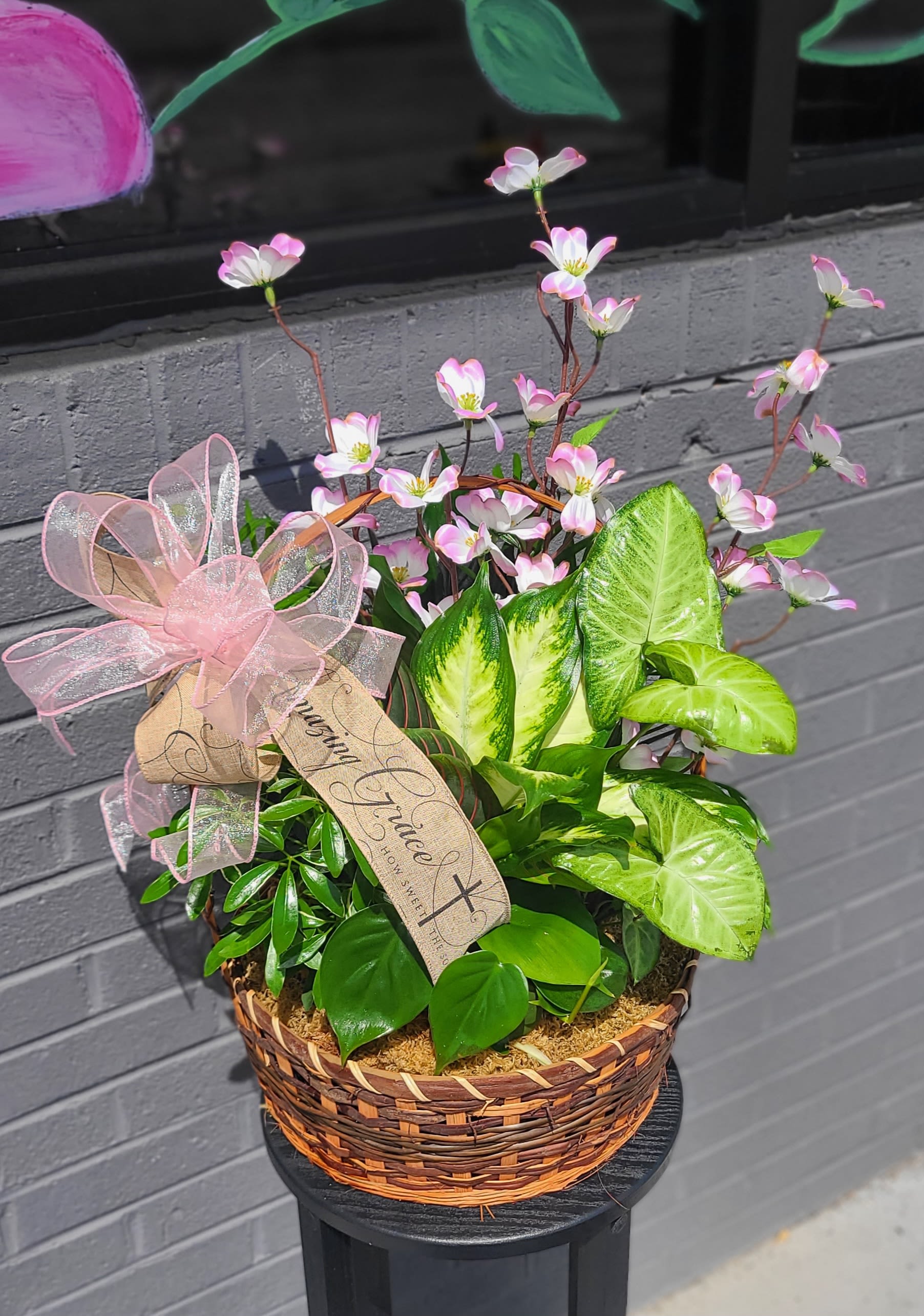 Garden Dish - Glossy green foliage tiny with bow and blooms that spill from a round wicker basket to create a living plant gift perfect for a housewarming or thinking-of-you moment. This mixed potted plant basket reads more like a lasting, cheerful present than a cut-flower bouquet with ribbon and decorative tag that adds a romantic, gift-ready touch that makes the recipient feel seen.  Several leafy houseplants with varied textures-broad variegated leaves, smaller rounded foliage, and upright stems topped with delicate ribbon and blooms arranged over a moss-lined base inside a woven container. The basket is finished with a large ribbon bow and hanging tag; moss covers the soil for a tidy, natural look Ideal for tabletops, entryways, or brightening a kitchen windowsill. This composition brings durable greenery and gentle floral accents into the home.  A thoughtful choice for birthdays, new homes, or a "just because" surprise, this living basket offers long-lasting enjoyment that keeps on giving as the plants grow. Water when the top inch of soil feels dry and place in bright, indirect light; the compact woven basket and decorative ribbon make it ready to present as soon as it arrives.`