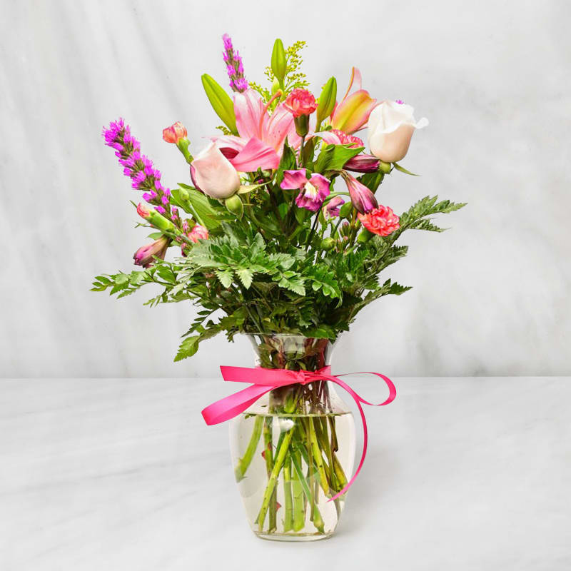 Mixed bouquet of pink lilies, roses, and carnations in a clear glass vase with a pink ribbon