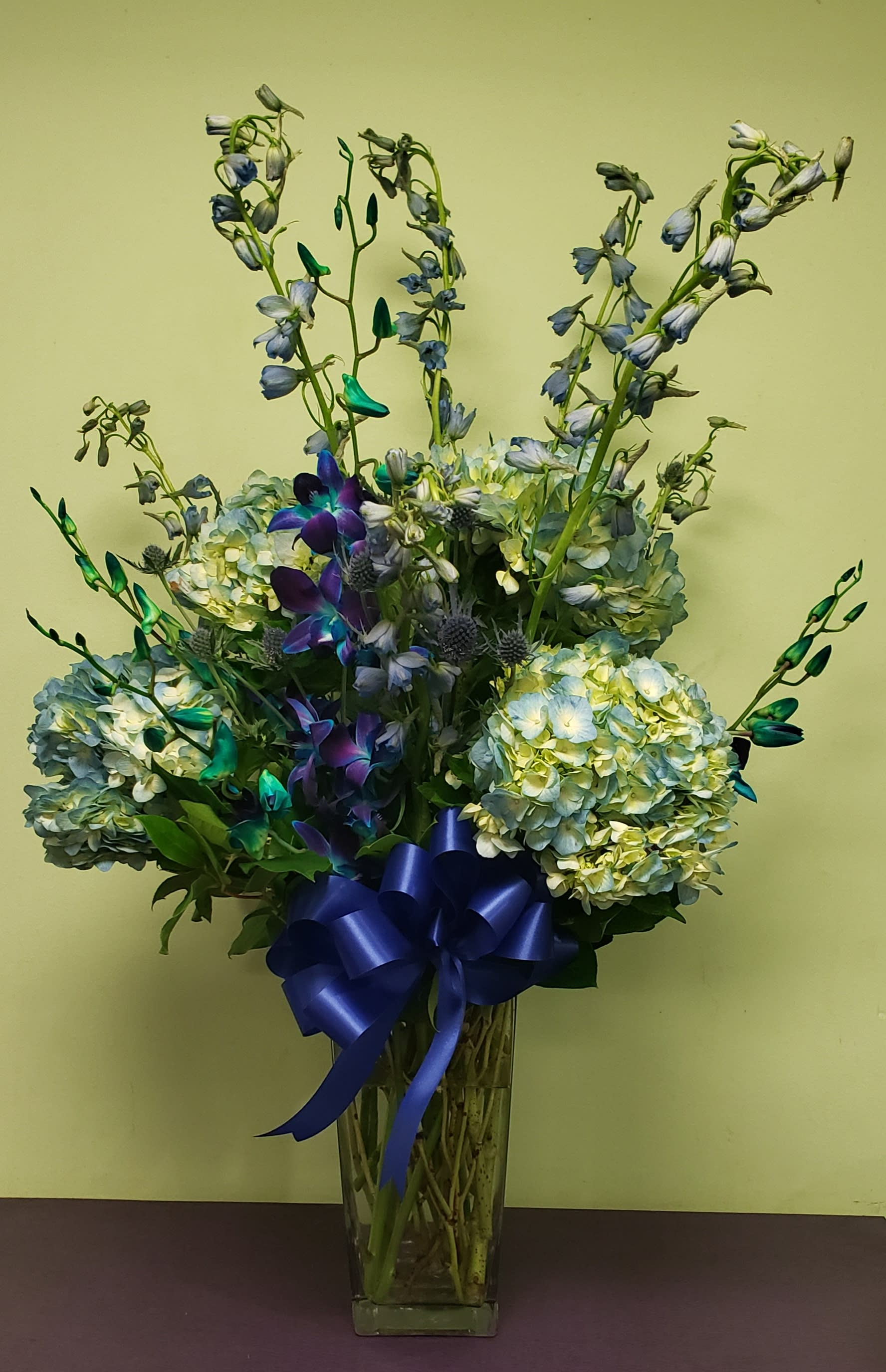 Blue hydrangea and orchid arrangement in a tall glass vase with a blue ribbon