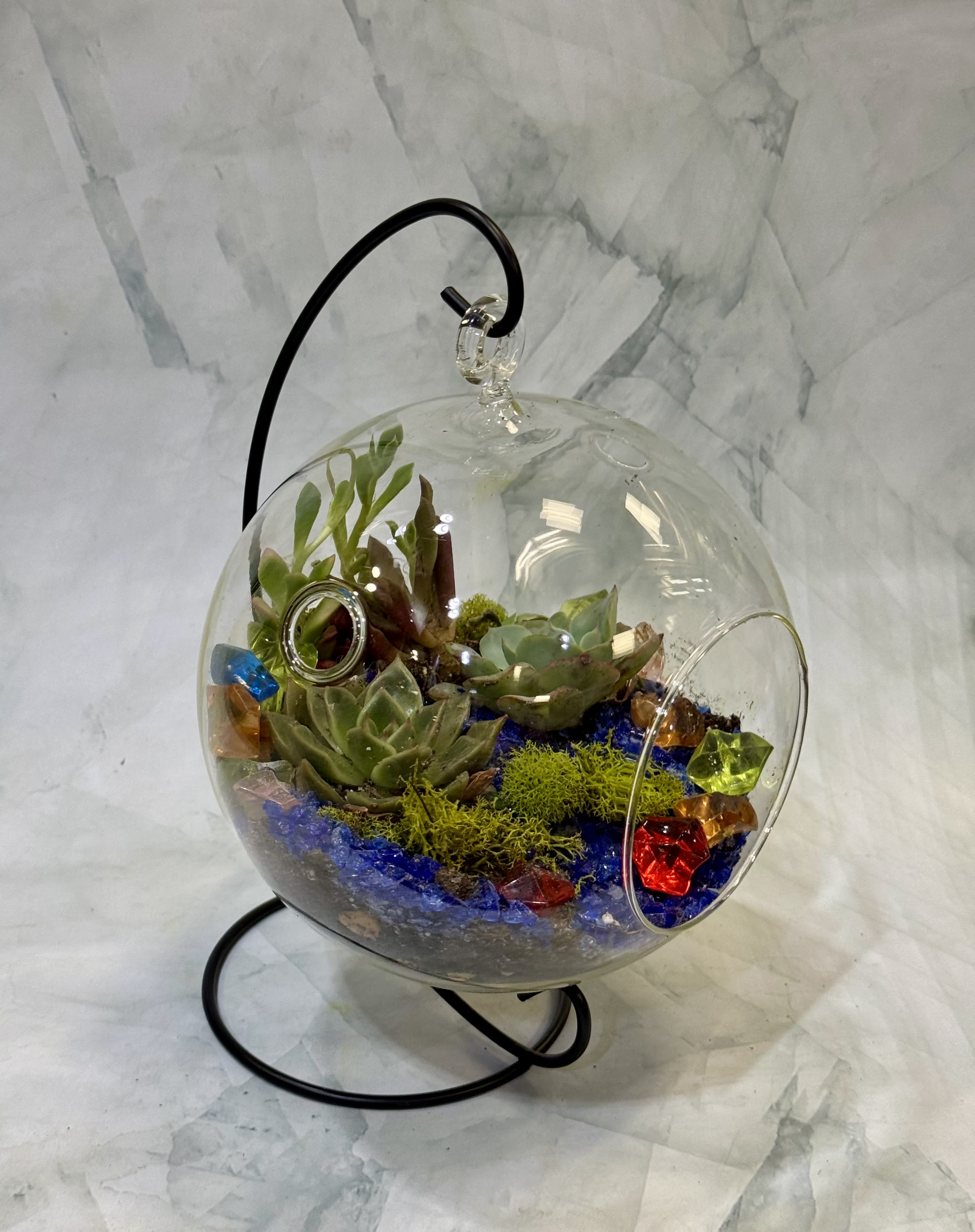 Glass globe terrarium on a black stand with green succulents, blue stones, moss, and colorful glass gems