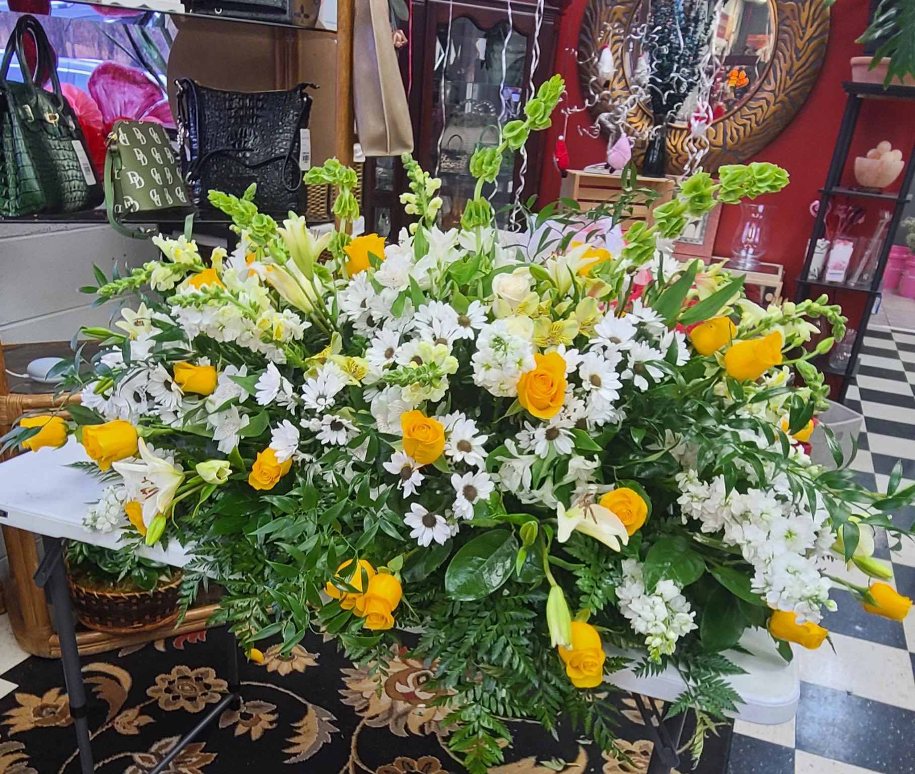 Casket Spray Loving the Daisy - A large, elongated sympathy spray in sunny yellow and soft white that gives a warm, hopeful presence at a funeral or memorial service. This casket-style or altar piece centers on bright yellow roses paired with white lilies and clusters of daisies, the combination creating a comforting visual that reads as both uplifting and gentle for the person you're honoring.  Densely massed blooms-white stock and chrysanthemums for soft texture, spired snapdragons for vertical variety, and tall green Bells of Ireland for structured accents-are arranged horizontally in a full, low profile. The closely packed flowers form a lush, cascading silhouette framed by rich greenery, so the arrangement reads beautifully from the front and across the length of a casket or display table.  Made at a large scale to suit funeral and memorial settings, this spray is designed to be immediately impactful without overpowering the space. It's the kind of tribute that lets the recipient's family feel seen and comforted: warm yellow for remembrance and white for peace. Please contact us for delivery timing, placement options, or size adjustments to ensure it fits the service and setting you have in mind.
