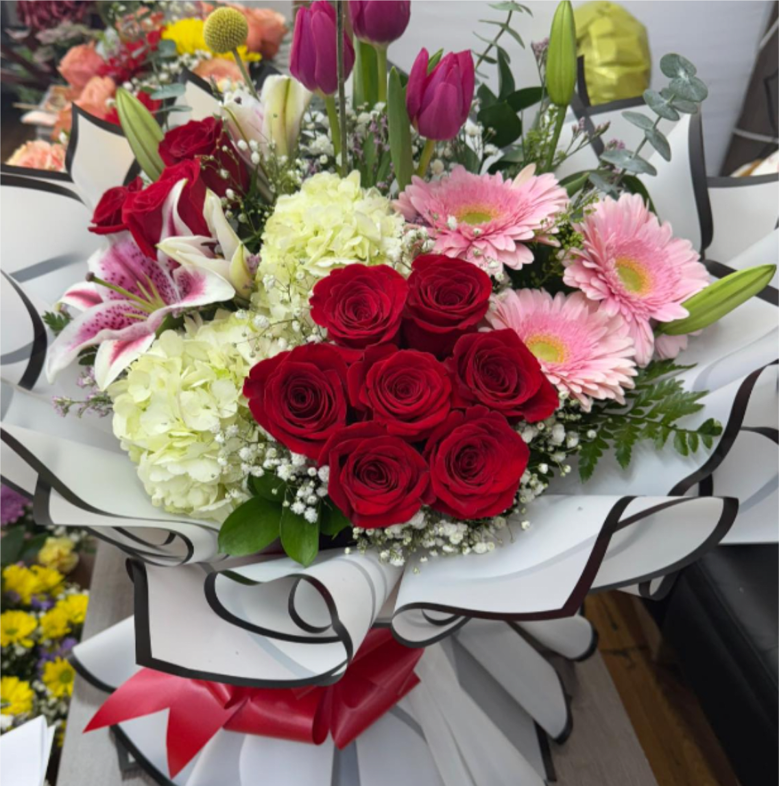 True Love Bouquet - A lush cluster of velvety red roses and soft pink gerbera daisies sets a romantic, celebratory tone for this anniversary roses bouquet - the kind of gift that makes the person you're thinking of feel cherished. Pale green hydrangea and white lilies add volume and a gentle fragrance, while tall purple tulips (in select arrangements) lift the composition for a contemporary silhouette suited to birthdays, Valentine's Day, or any special milestone.  The mixed textures - dense rose heads, ruffled hydrangea, open daisy faces, and lily buds - create a full, layered look that reads as both luxurious and joyful. Baby's breath fills the spaces with an airy softness and the stems are wrapped in large white paper with black edging, finished with a red ribbon for a polished, gift-ready presentation. Colors include red, pink, white, green, and purple for a vibrant, romantic palette.  This is an ideal choice when you want her to feel truly celebrated - for anniversaries, birthdays, or a heartfelt "I love you." Sizes and stem counts vary by selection; arrange for same-day delivery when available and trim stems and refresh water on arrival to extend vase life.