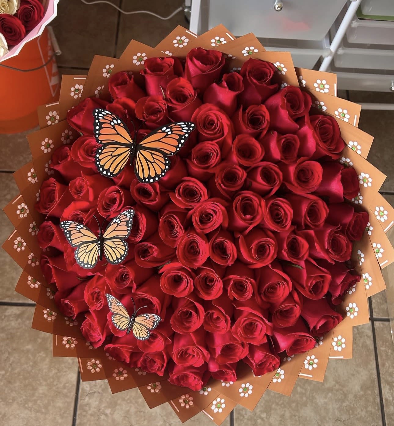 The Barro Ramo Buchón - A tightly packed, round bouquet of red roses that reads pure romance at first glance-perfect for anniversaries, Valentine's Day, or any love-and-romance moment when you want the person you're thinking of to feel deeply cherished. The dense, buchón-style shape creates a bold, celebratory silhouette that says "I adore you" with rich color and floral intensity; it also makes a beautiful, heartfelt Mother's Day gift that feels special and unmistakable.  Every bloom is arranged close together for a lush, velvety surface of crimson petals, framed by layered orange-brown decorative paper printed with tiny floral motifs for warm contrast and texture. Three delicate butterfly picks float across the front, adding a playful, whimsical touch to the classic rose statement without distracting from the bouquet's romantic impact.  Ideal for marking anniversaries, engagements, or a meaningful surprise, this arrangement sends a clear message of love and devotion to the recipient. The wrapped presentation is ready to carry and display; keep the stems in fresh water, trim a half inch off the ends before arranging in a vase, and replace water daily to prolong the life of the roses.