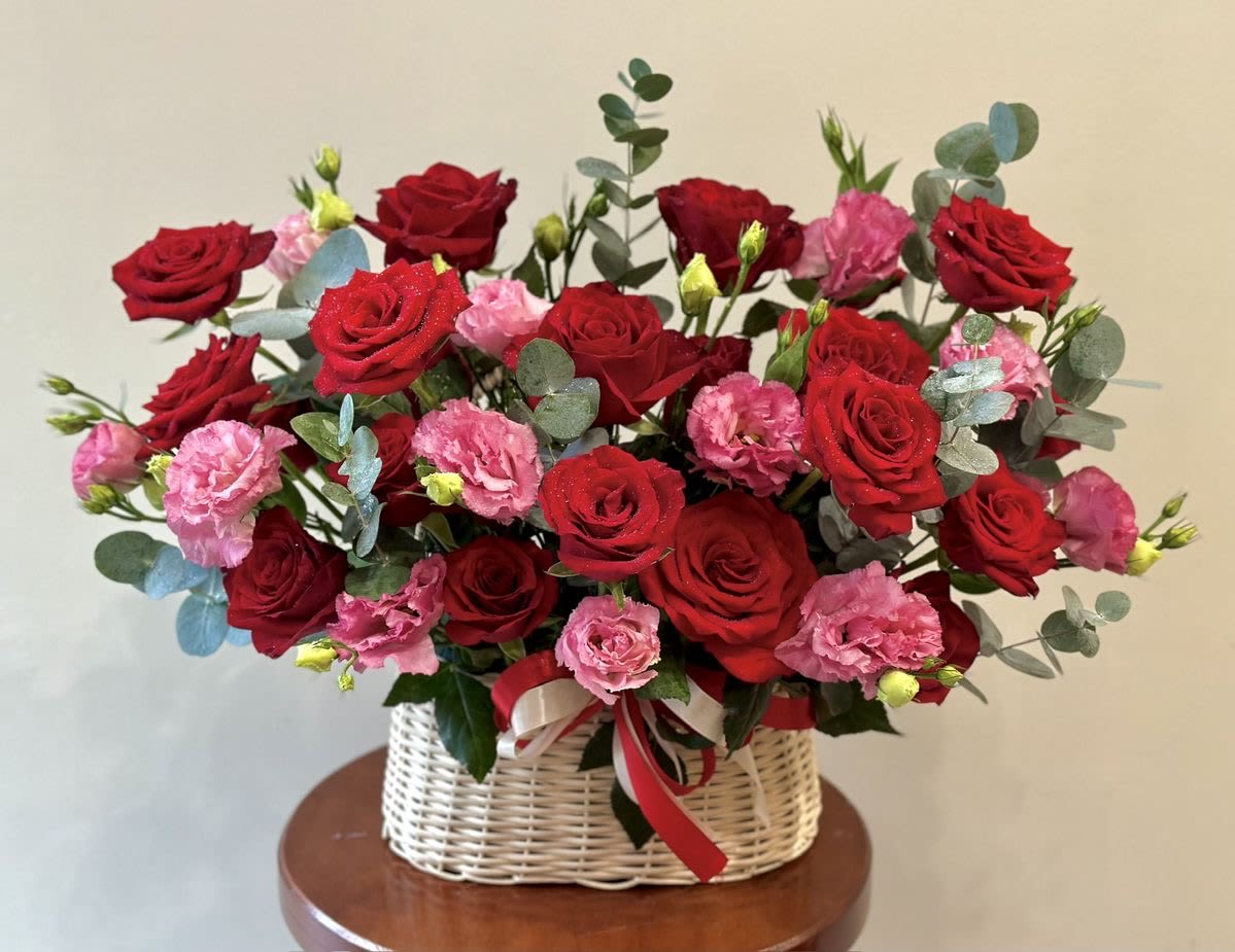 Un jardin de rosas rojas  - A lush cascade of red roses and soft pink carnations fills a low woven basket, creating a romantic birthday bouquet or anniversary arrangement that reads as both elegant and approachable. Un jardin de rosas rojas is designed to make the person you're thinking of feel adored-deep crimson roses convey passion while the blushing pink carnations soften the mood into warmth and tenderness, perfect for Valentine's Day, anniversaries, or a heartfelt "just because" romantic gift.  Blue-green eucalyptus weaves through the blooms, adding airy texture and a cool contrast to the warm red and pink palette; touches of white and fresh green foliage brighten the composition and keep it visually light. The arrangement sits low and spreading in a handwoven basket tied with a ribbon, so it looks ready to display on a dining table or desk without extra fuss. Sized to give an abundant presence without towering, it arrives gift-ready and easy to place where it will be seen-and felt-right away.