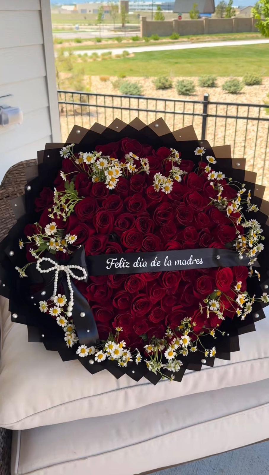 La mejor mama  - A dramatic, romantic bouquet of deep red roses mingled with delicate white daisies creates an unforgettable Mother's Day or anniversary gift that reads boldly from across the room. This large, tightly packed arrangement uses the contrast of velvety red petals against crisp white daisy centers and layered black wrapping to make the recipient feel both adored and deeply seen-perfect for the mother, partner, or special woman you want to honor.  The bouquet features abundant deep red roses accented by scattered small white daisies, all arranged close together for rich visual impact. Finished in layered black wrapping paper with a wide black ribbon across the front and a decorative pearl-like bow near the lower left, the presentation leans luxe and slightly dramatic while the daisies add a soft, tender touch. The black ribbon text in Spanish suggests a Mother's Day sentiment, making this an especially fitting choice for that occasion.  Choose this arrangement when you want someone to feel celebrated, cherished, and noticed-whether for Mother's Day, an anniversary, or a meaningful romantic gesture. As a large bouquet, it makes a strong centerpiece or focal gift; for best longevity, keep the stems in fresh water, recut stems before placing them in a vase, and refresh the water every two days to maintain bloom freshness.