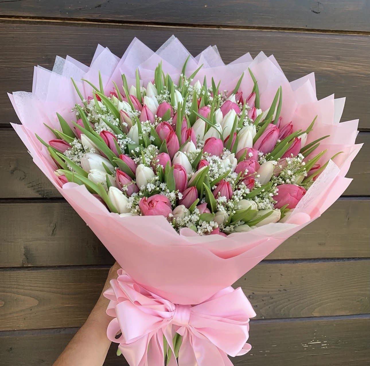 Tulip lovers choice - A lush cascade of pink and white tulips makes a joyful first impression-this flower arrangement is a tulip bouquet perfect for anniversaries, birthdays, or simply to let someone know you're thinking of them. The mix of blushing pinks and pure whites reads romantic and fresh, the kind of gift that will make the person you've chosen feel genuinely celebrated and seen.  Densely packed tulip heads are softened by small clusters of white filler blooms and long, pointed green leaves for contrast and texture. The stems are wrapped in layered pink paper and tied with a large pink satin ribbon, giving the bouquet a polished, spring-like presentation that's ready to hand to the recipient. Compact yet generous in volume, it arrives looking full and vibrant-an elegant, cheerful choice for any celebratory moment.